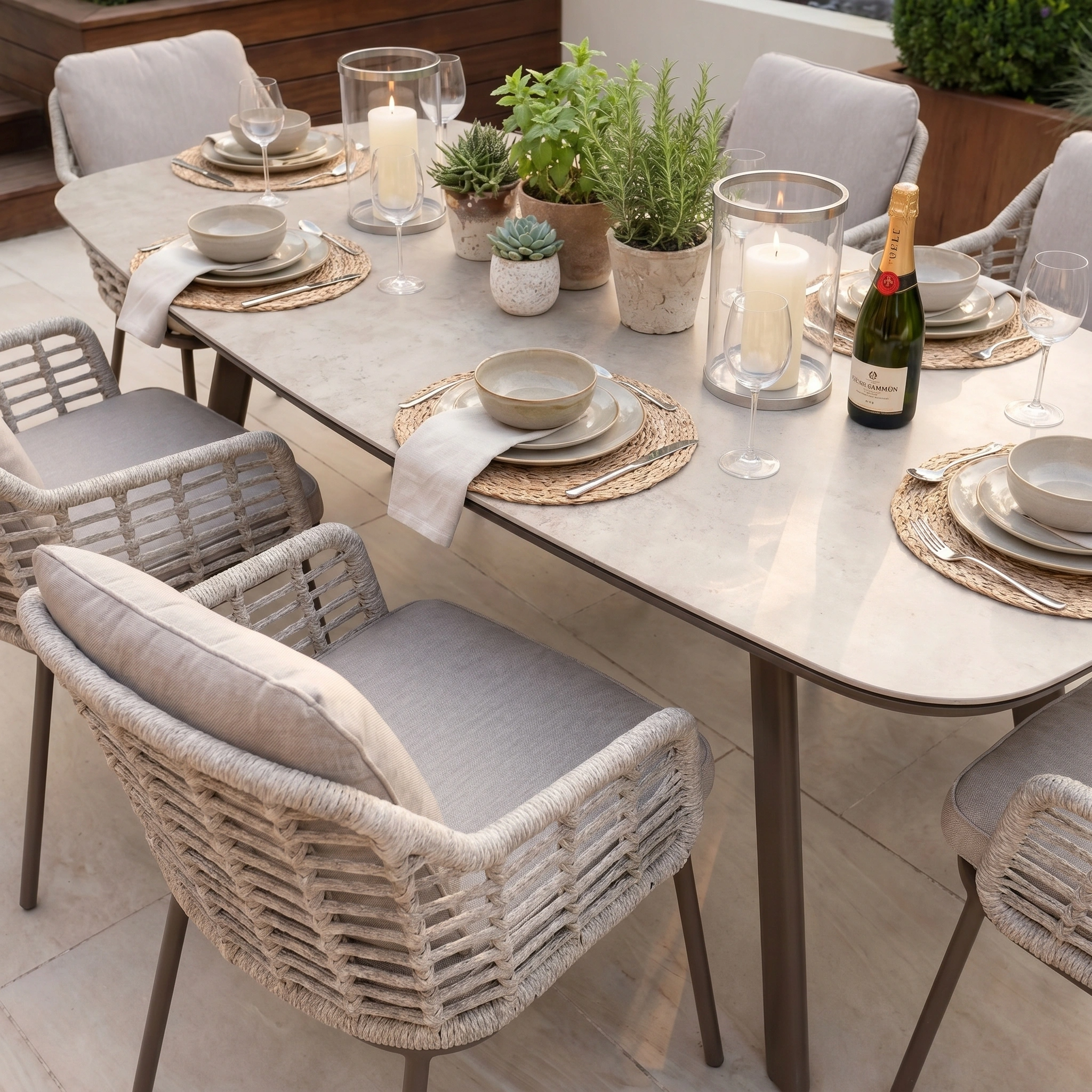 The Sutera 6 Seat Curved Rectangular Dining Set in Putty is elegantly styled outdoors with plates, glasses, candles, plants, and champagne—ideal for gatherings around this modern six-seat set.