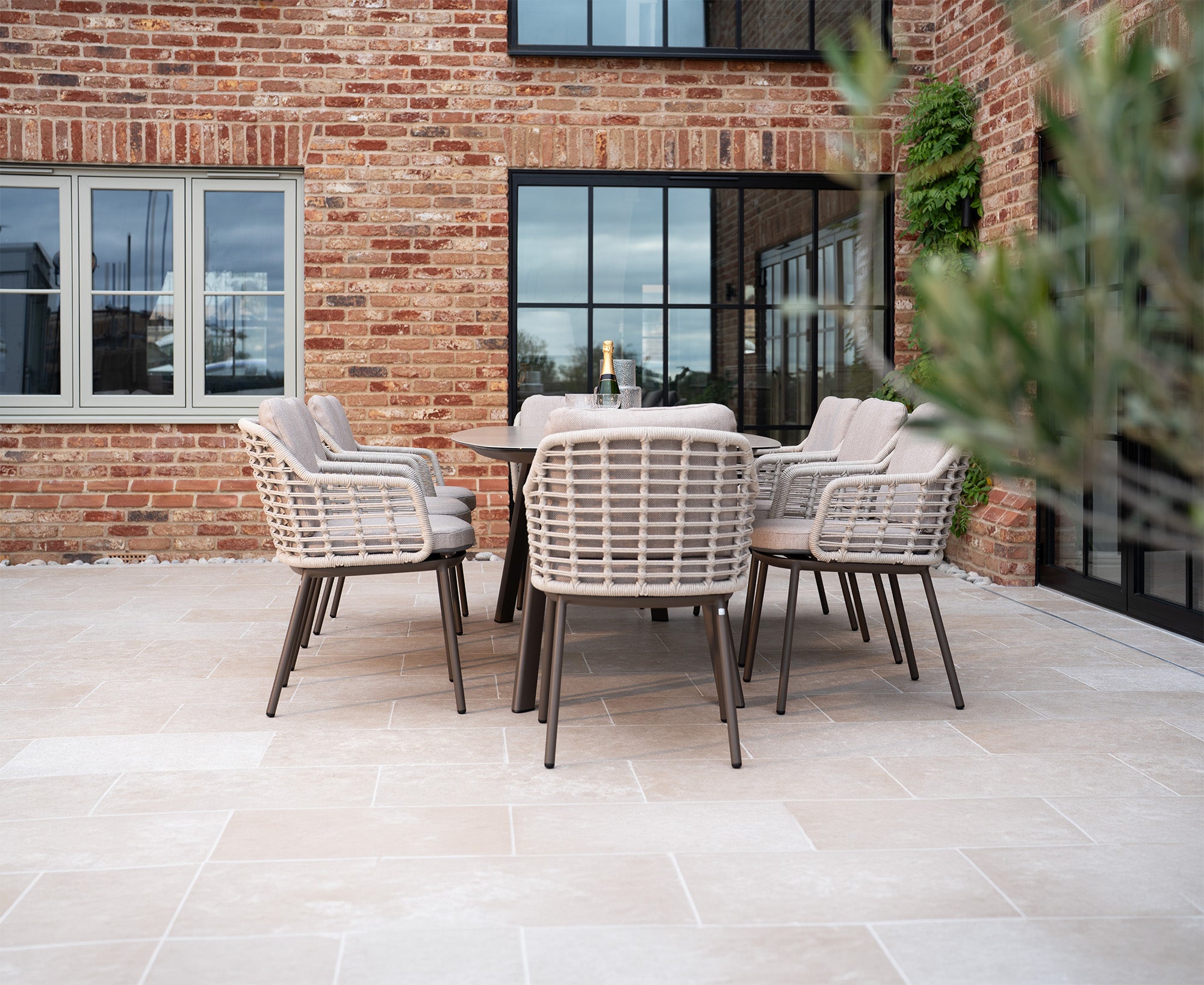 Enjoy your outdoor patio with the Ali testing metafield set, featuring six woven chairs arranged around a table with a sturdy aluminium frame, all beautifully positioned in front of a brick building with expansive windows.