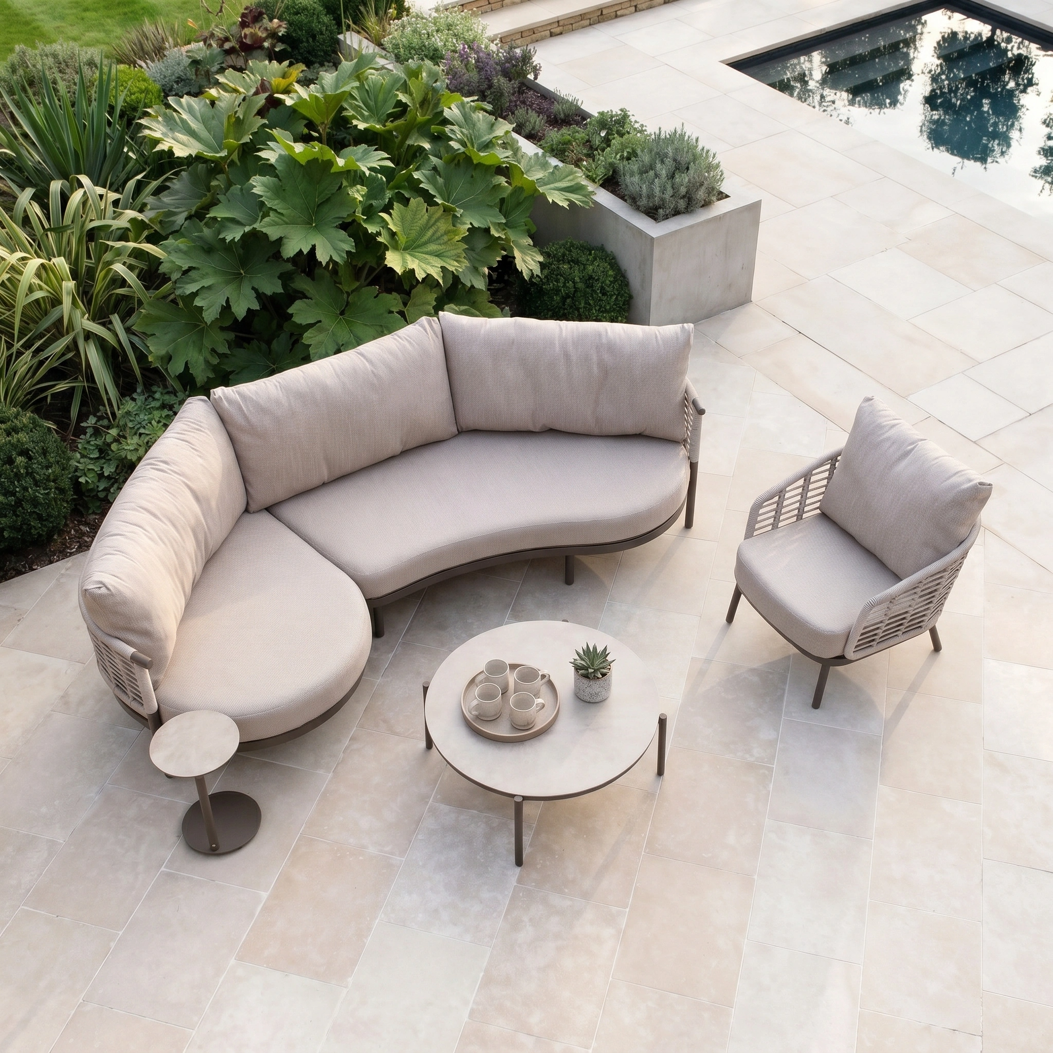 Modern outdoor patio with the Sutera Curved Chaise Sofa Set with Armchair in Putty, round tables, and all-weather cushions amid lush plants by a pool on light stone tiles.