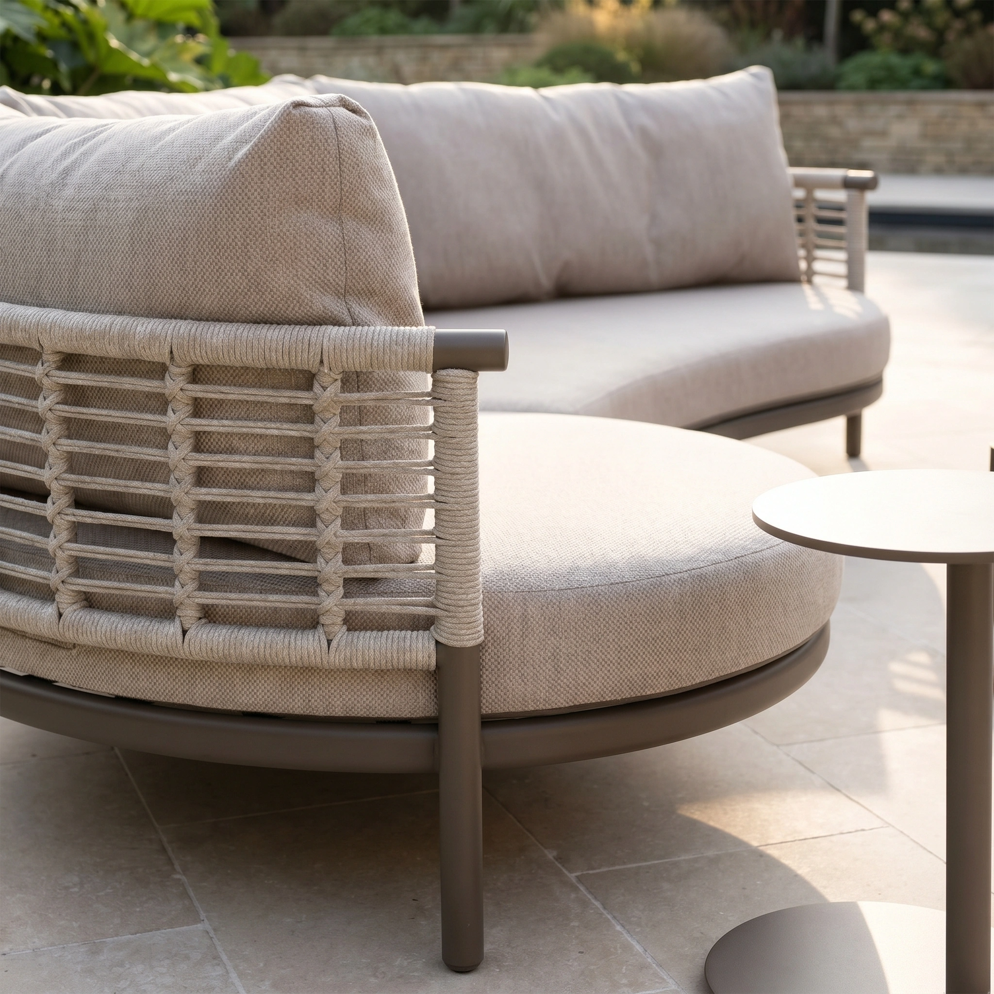 The Sutera Curved Chaise Sofa Set with Armchair in Putty features all-weather cushions and a woven backrest, arranged beside a small round table on a stone patio.
