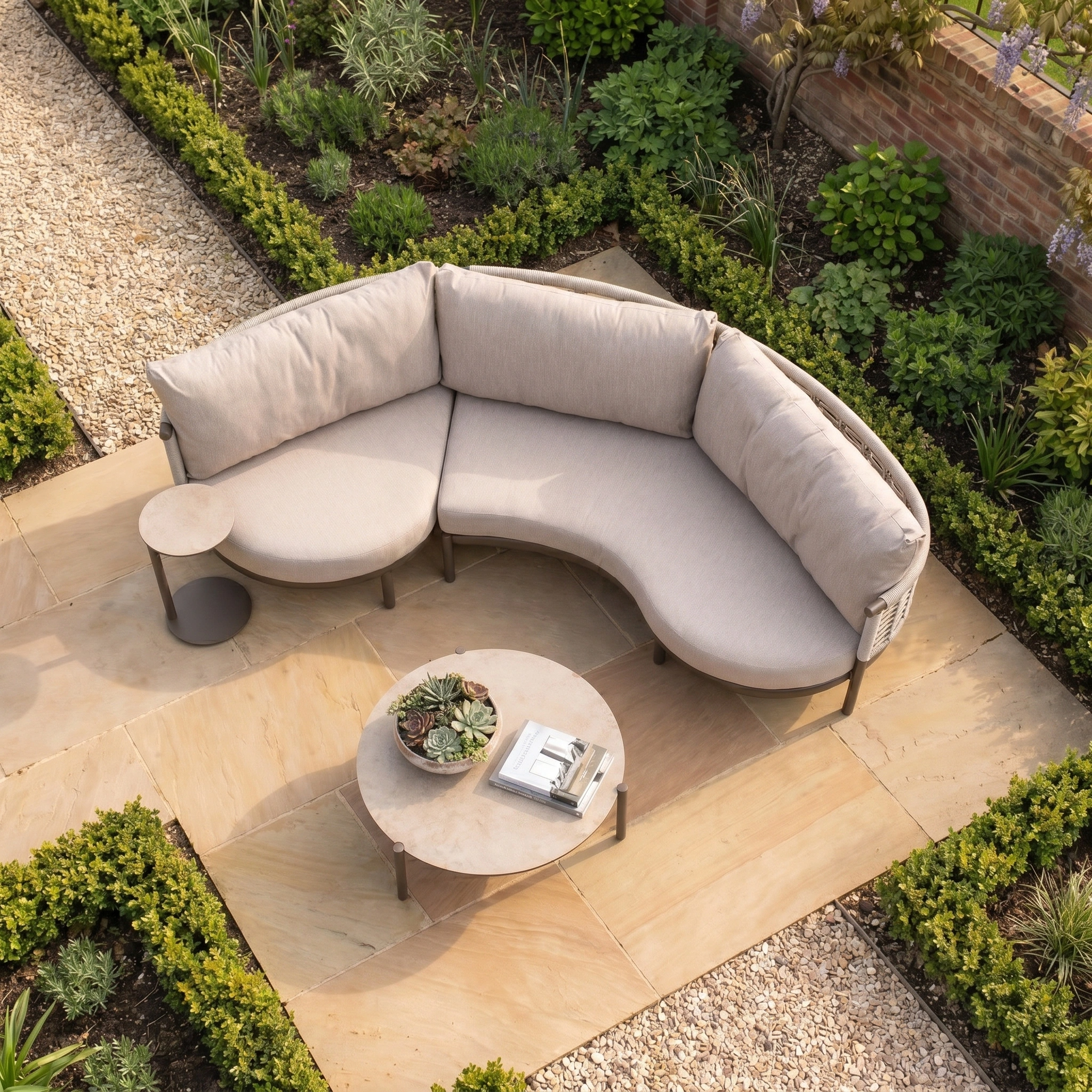 The Sutera Curved Chaise Sofa Set in Putty features beige woven rope design, round tables, and all-weather cushions, set on a stone patio amid lush green garden plants.