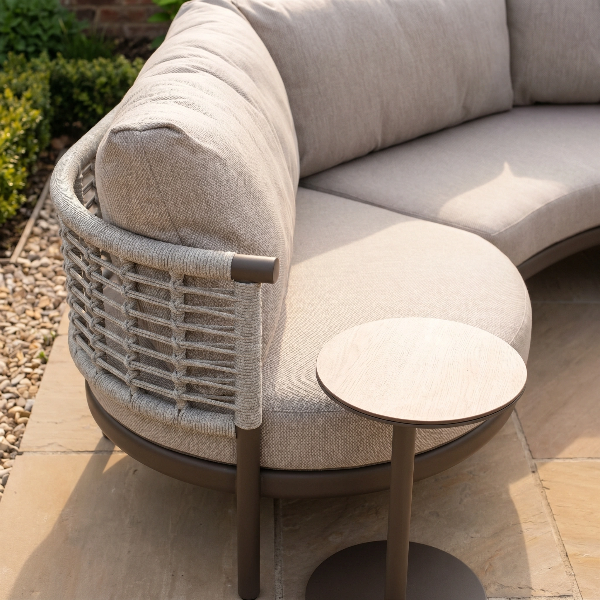 The Sutera Curved Chaise Sofa Set in Putty features woven rope and light beige all-weather cushions, placed beside a small round wooden side table on a stone patio.