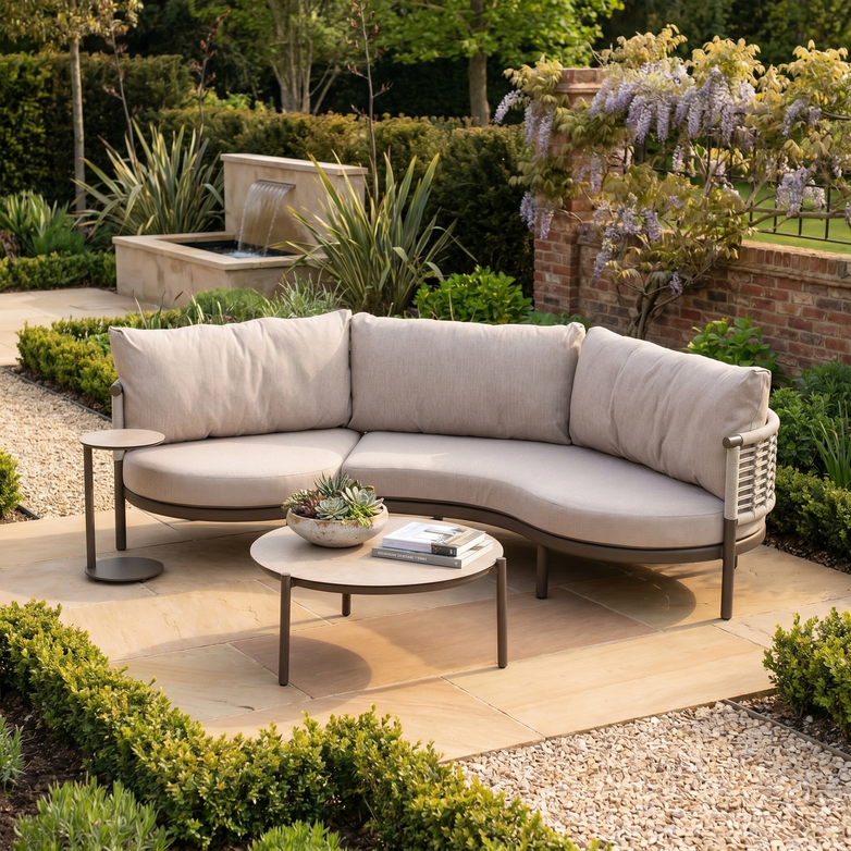 The Sutera Curved Chaise Sofa Set in Putty, featuring all-weather cushions and a round table, creates a cozy retreat in a garden surrounded by lush plants and vibrant flowers.