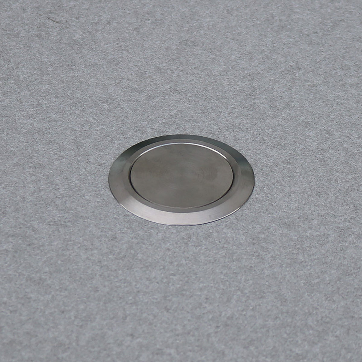 A round metal disk embedded in a flat, light gray surface, reminiscent of the sleek finishes seen on the Vela Corner Sofa Set in Wheat and similar contemporary outdoor seating arrangements.