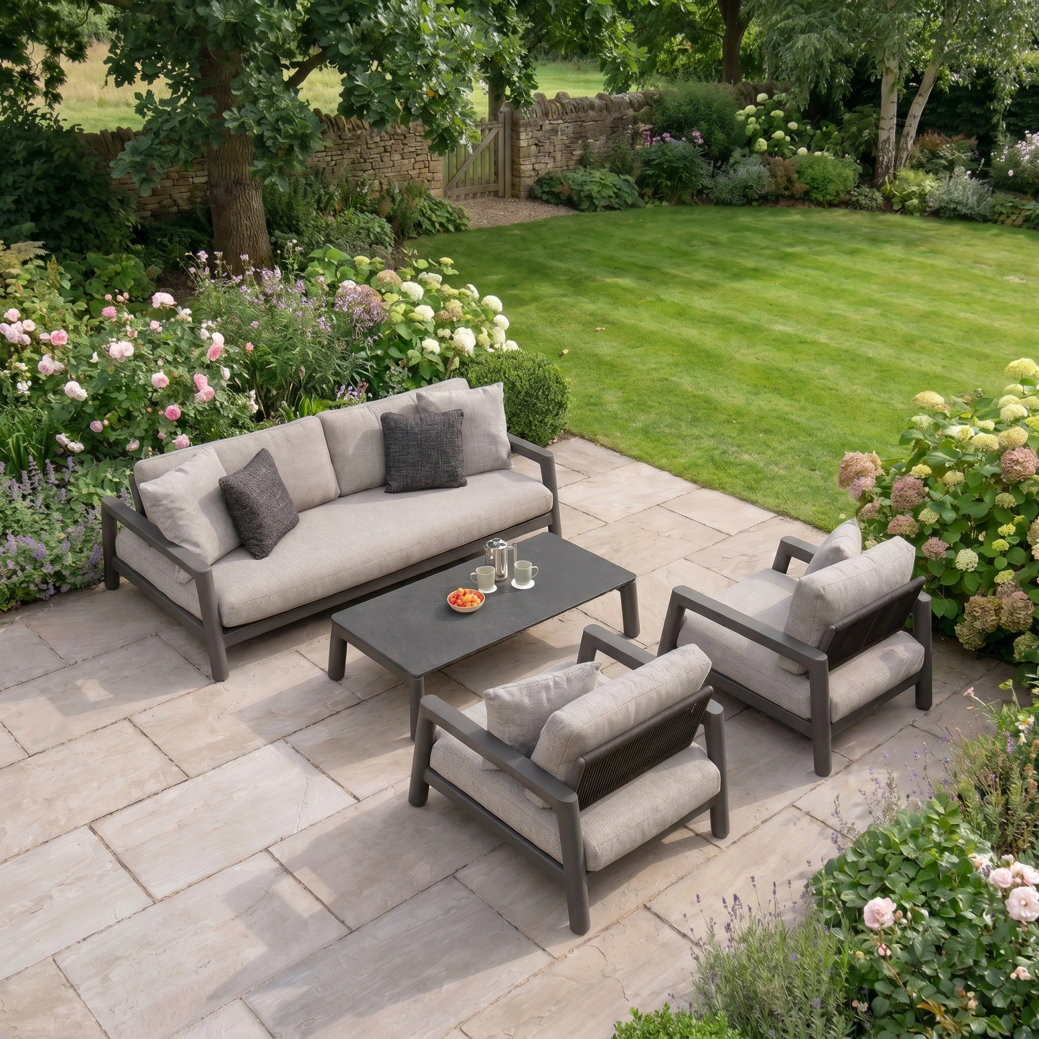 A lush garden frames the outdoor patio, furnished with Theia 3 Seat Sofa Set in Truffle—a gray cushioned modular sofa, two chairs, and a table topped with drinks and snacks.