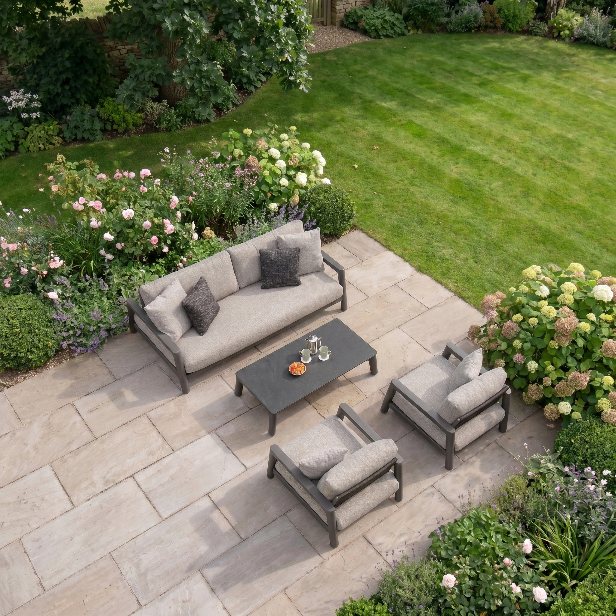 Patio featuring the Theia 3 Seat Sofa Set in Truffle—stylish modular seating and table, surrounded by a blooming garden and green lawn—perfect for relaxing with elegant outdoor furniture.