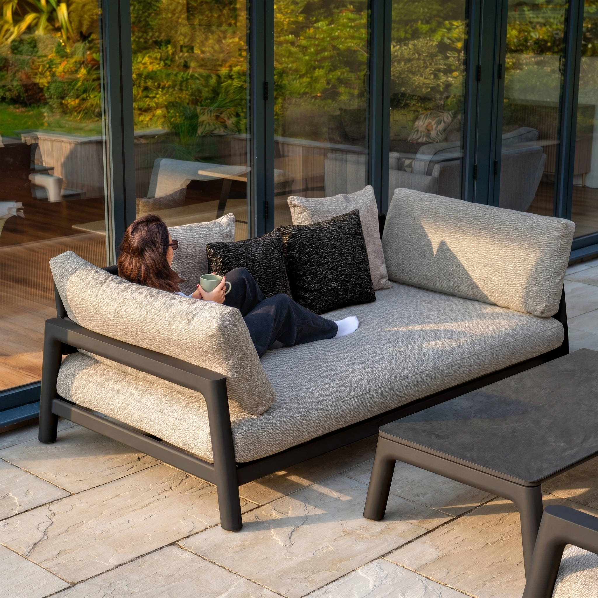 A person relaxes with a mug on the Theia 3 Seat Sofa Set in Truffle by glass doors, enjoying the garden view.