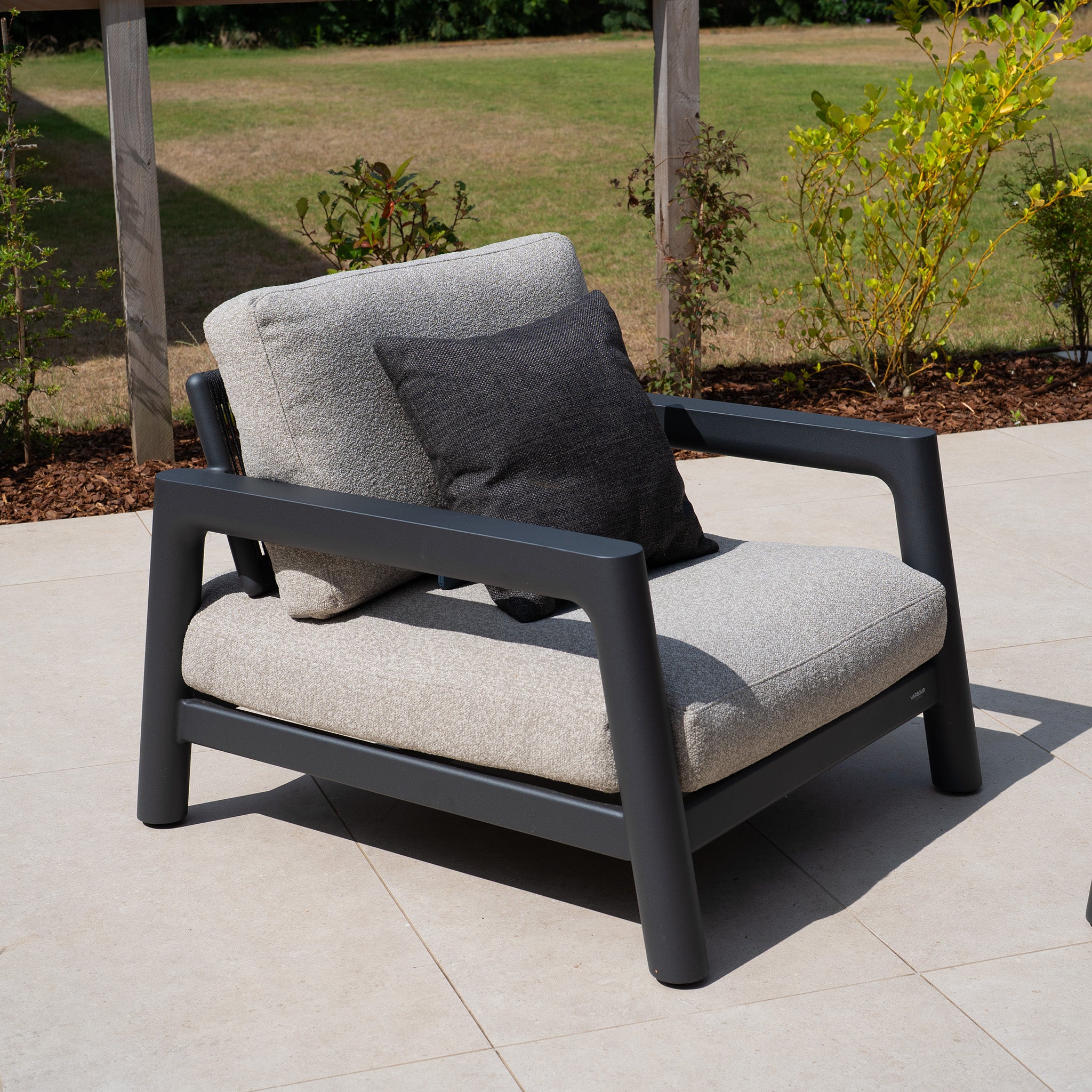 Gray outdoor armchair with light cushions and a dark pillow, placed on a tiled patio with greenery behind—perfect for complementing your outdoor furniture collection or pairing with the Theia 3 Seat Sofa Set in Truffle from our modular range.