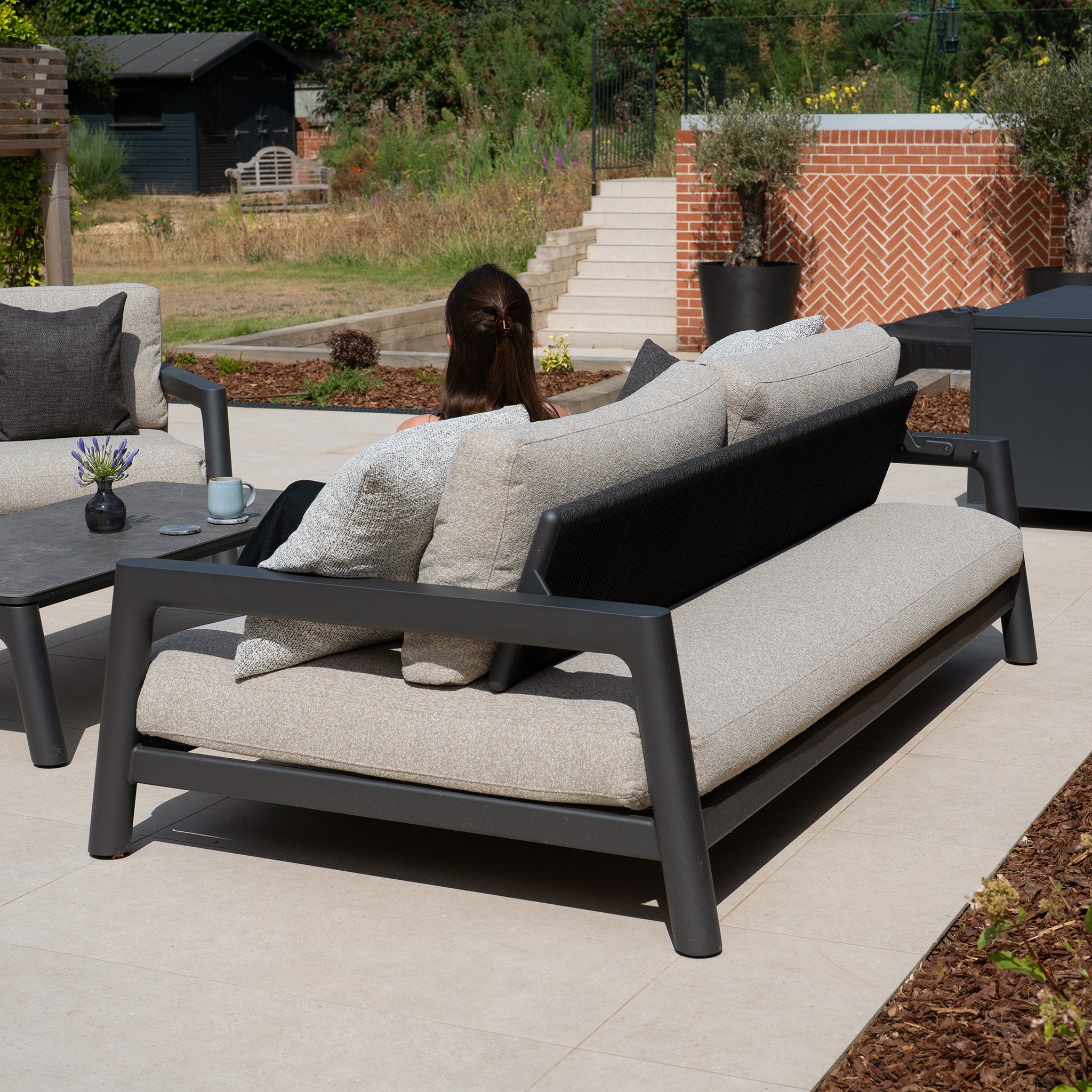 A woman sits on the Theia 3 Seat Sofa Set in Truffle with light cushions in a patio garden, facing away from the camera.