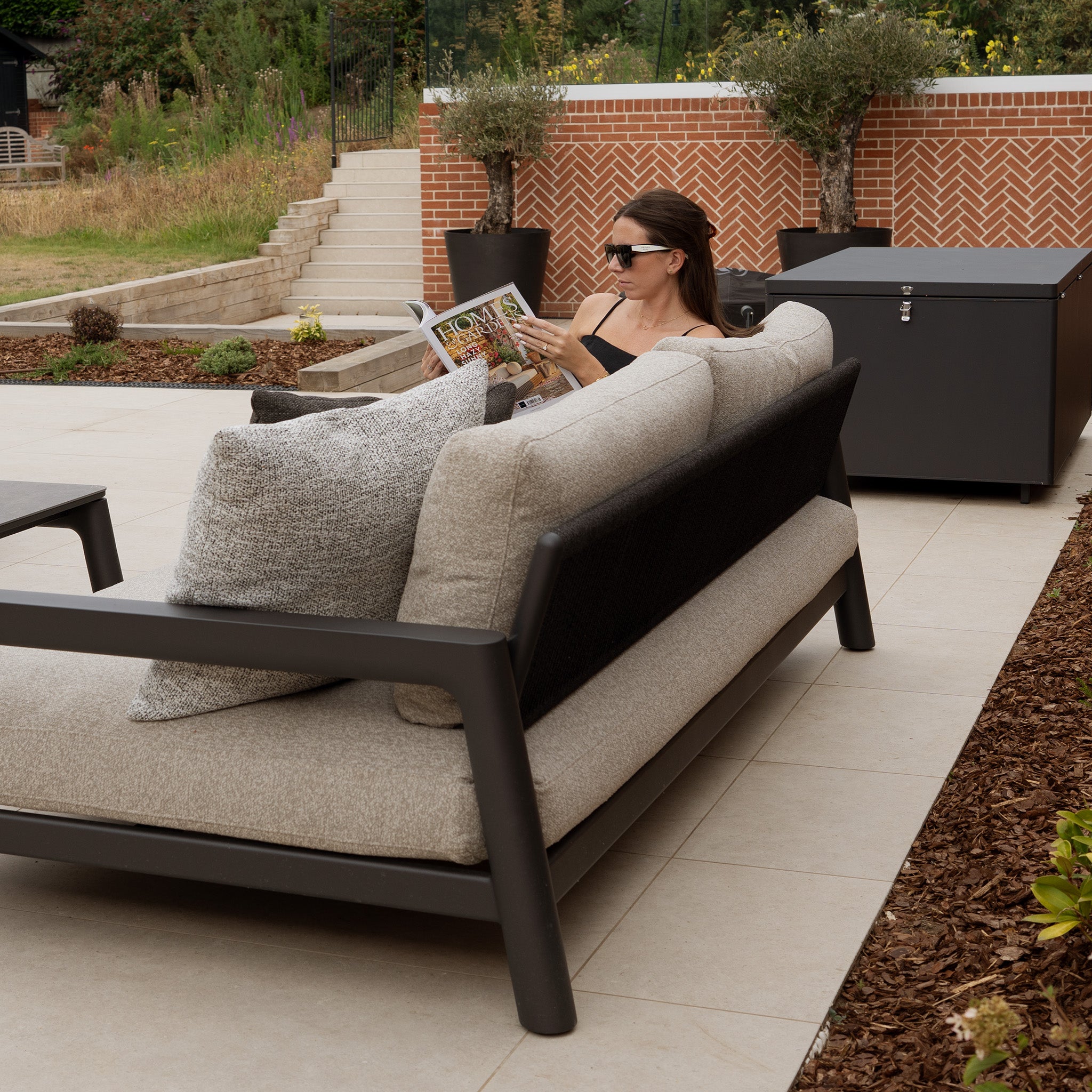 A woman in sunglasses reads a magazine on the Theia 3 Seat Sofa Set in Truffle, elegantly displayed in a garden setting and showcasing stylish outdoor furniture.