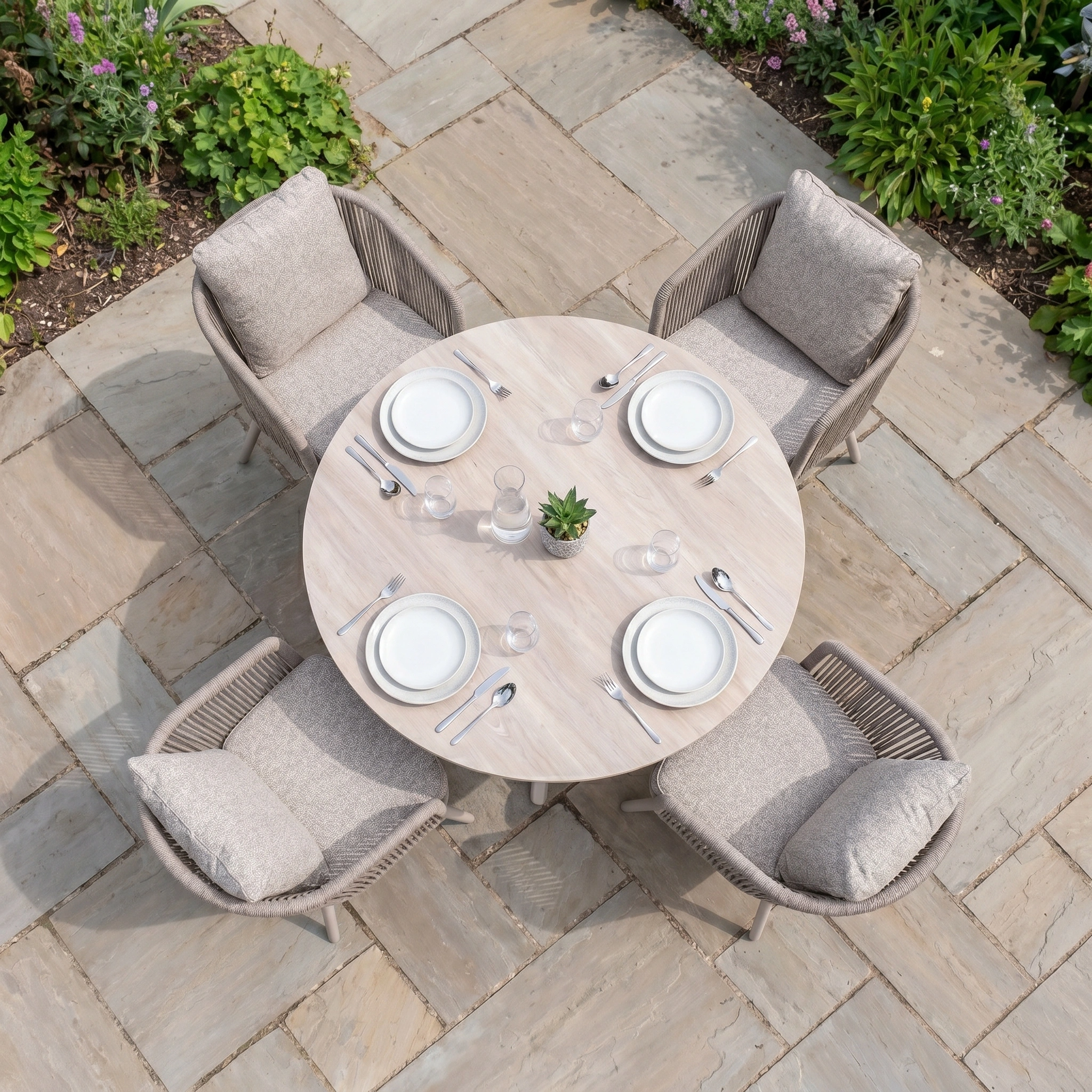 A round outdoor dining set for four with plates and cutlery, featuring cushioned chairs on a stone patio—beautifully styled as the Theia 4 Seat Round Rope Dining Set with Albatross Table in Oatmeal.