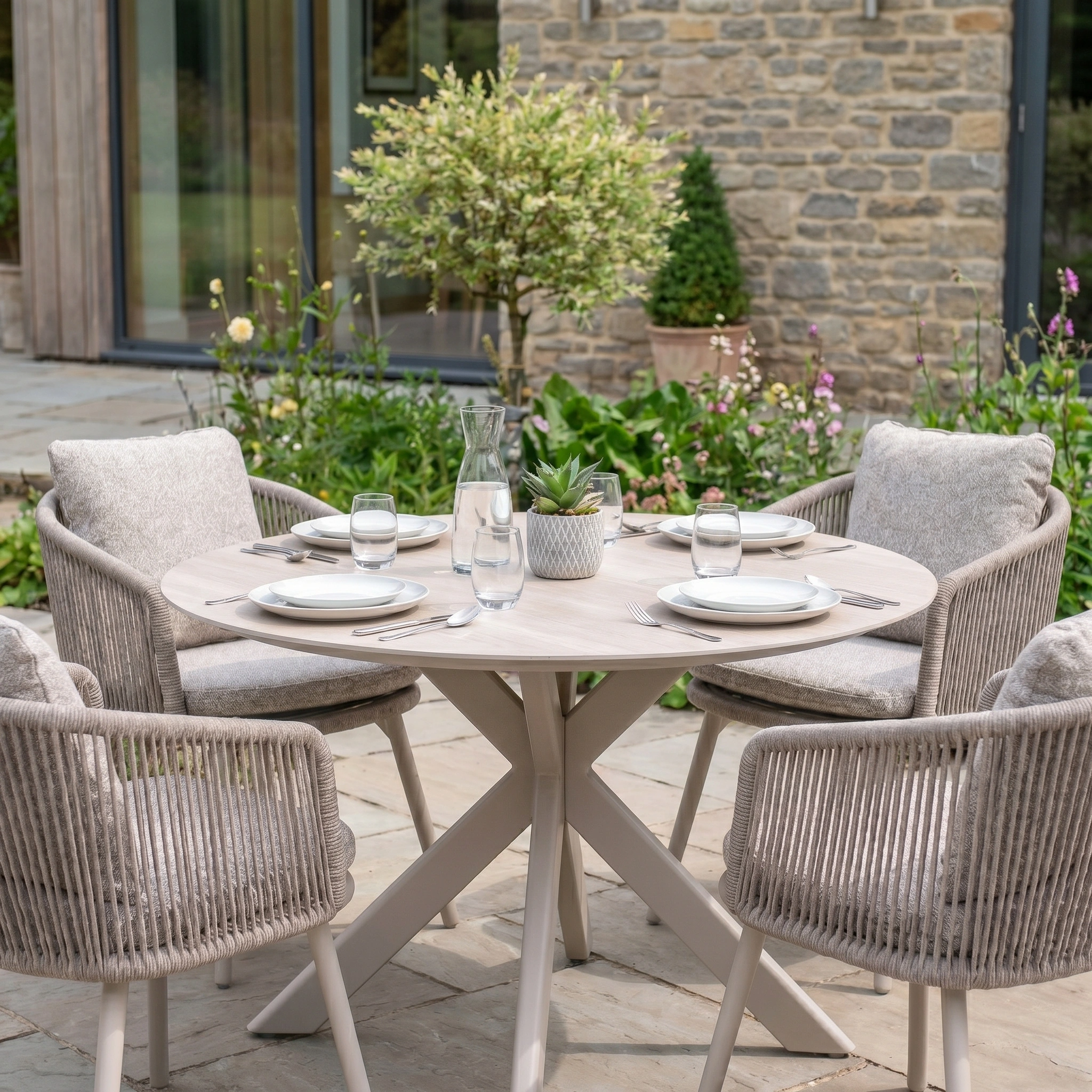 Theia 4 Seat Round Rope Dining Set with Albatross Table in Oatmeal includes a round patio table for four, cushioned chairs, stylish glassware, and a potted plant centerpiece.