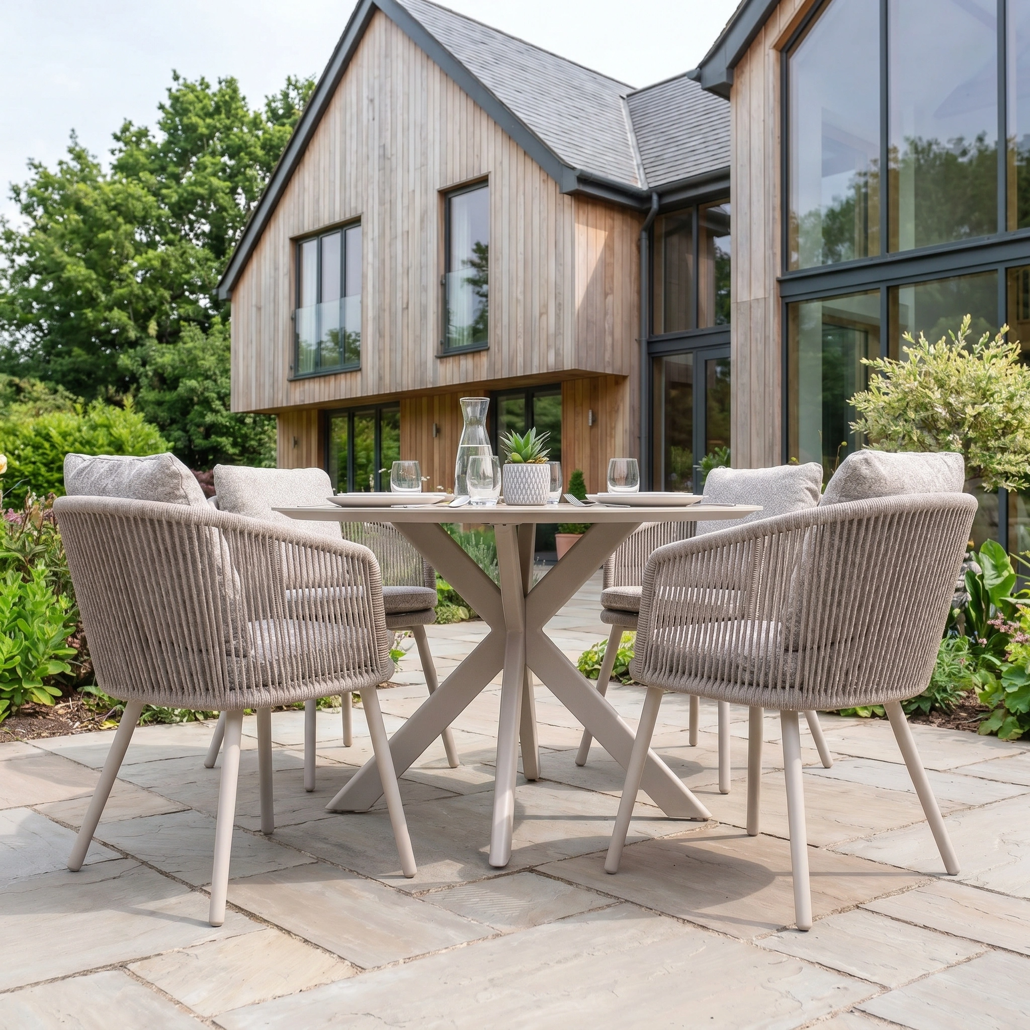 The Theia 4 Seat Round Rope Dining Set with Albatross Table in Oatmeal features four chairs and a round table, perfect for outdoor dining on a stone patio next to a wooden house.