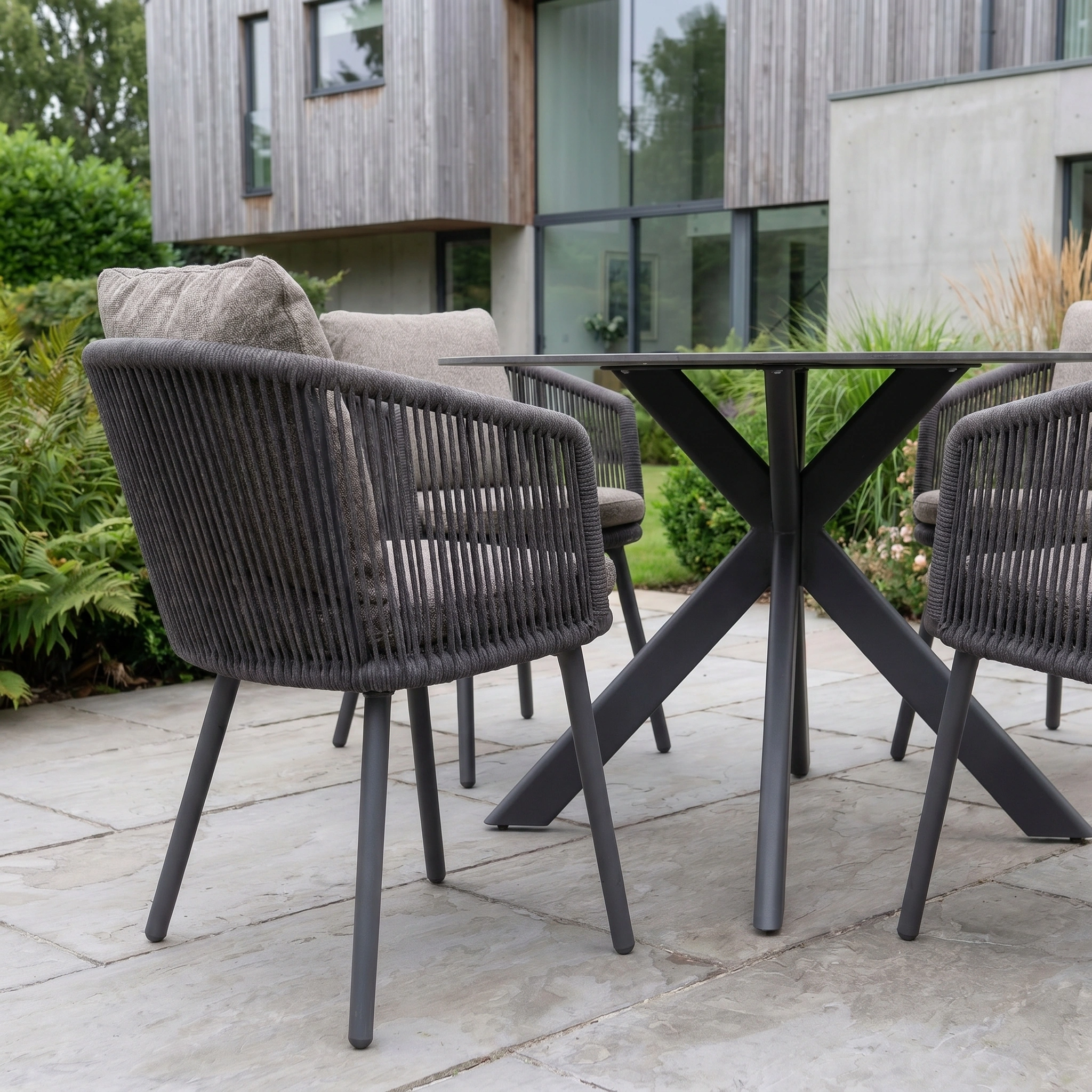 Theia 4 Seat Round Rope Dining Set with Albatross Table in Truffle styled on a stone terrace beside a modern house.