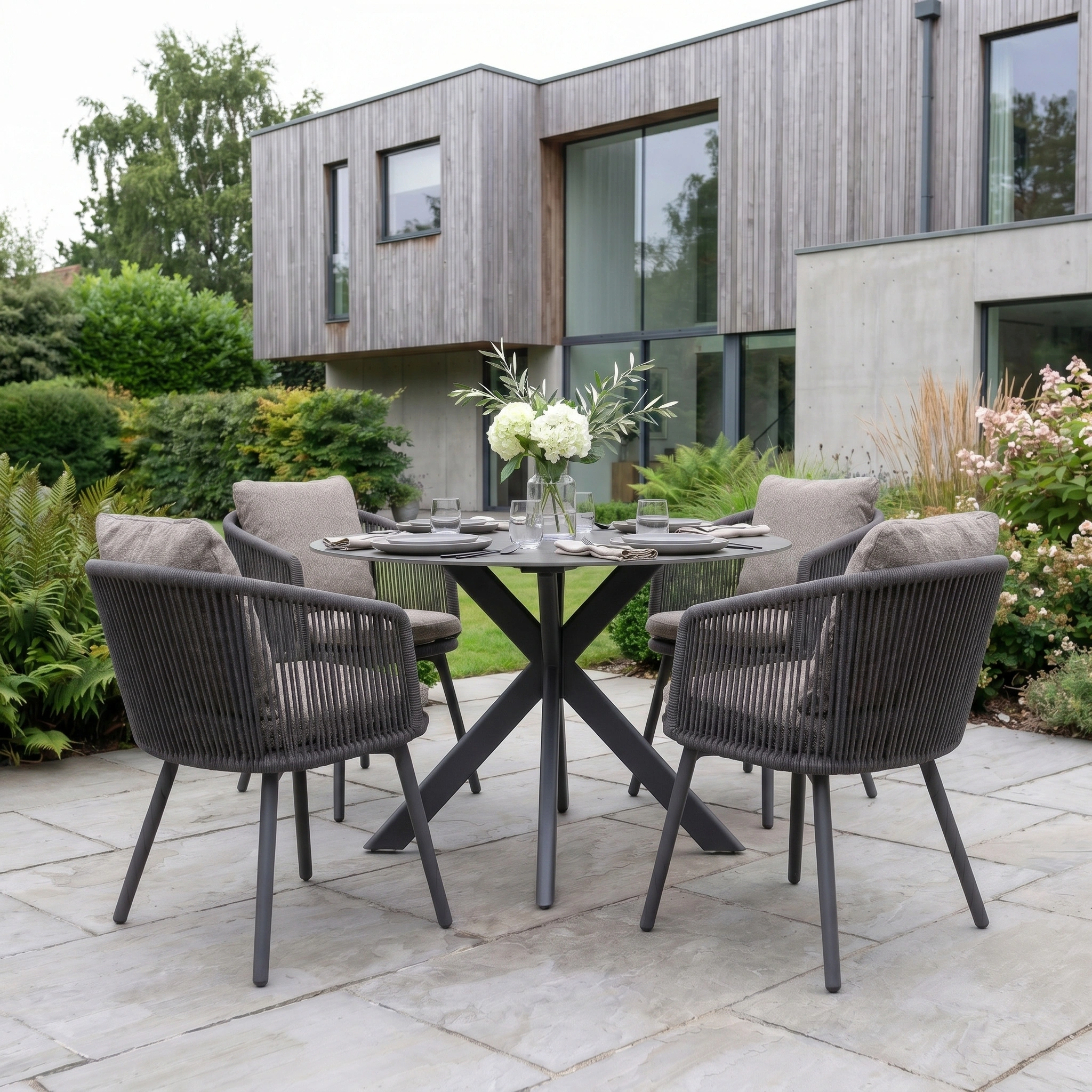 Theia 4 Seat Round Rope Dining Set with Albatross Table in Truffle, featuring four chairs and a round table, displayed on a stone patio before a modern house.