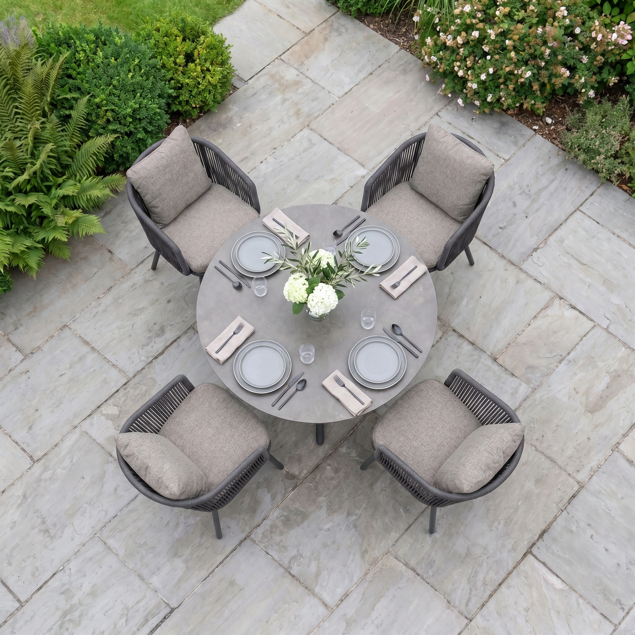 Theia 4 Seat Round Rope Dining Set with Albatross Table in Truffle—includes a round table and cushioned chairs, set with plates, napkins, and flowers on a stone patio—perfect for stylish outdoor dining.