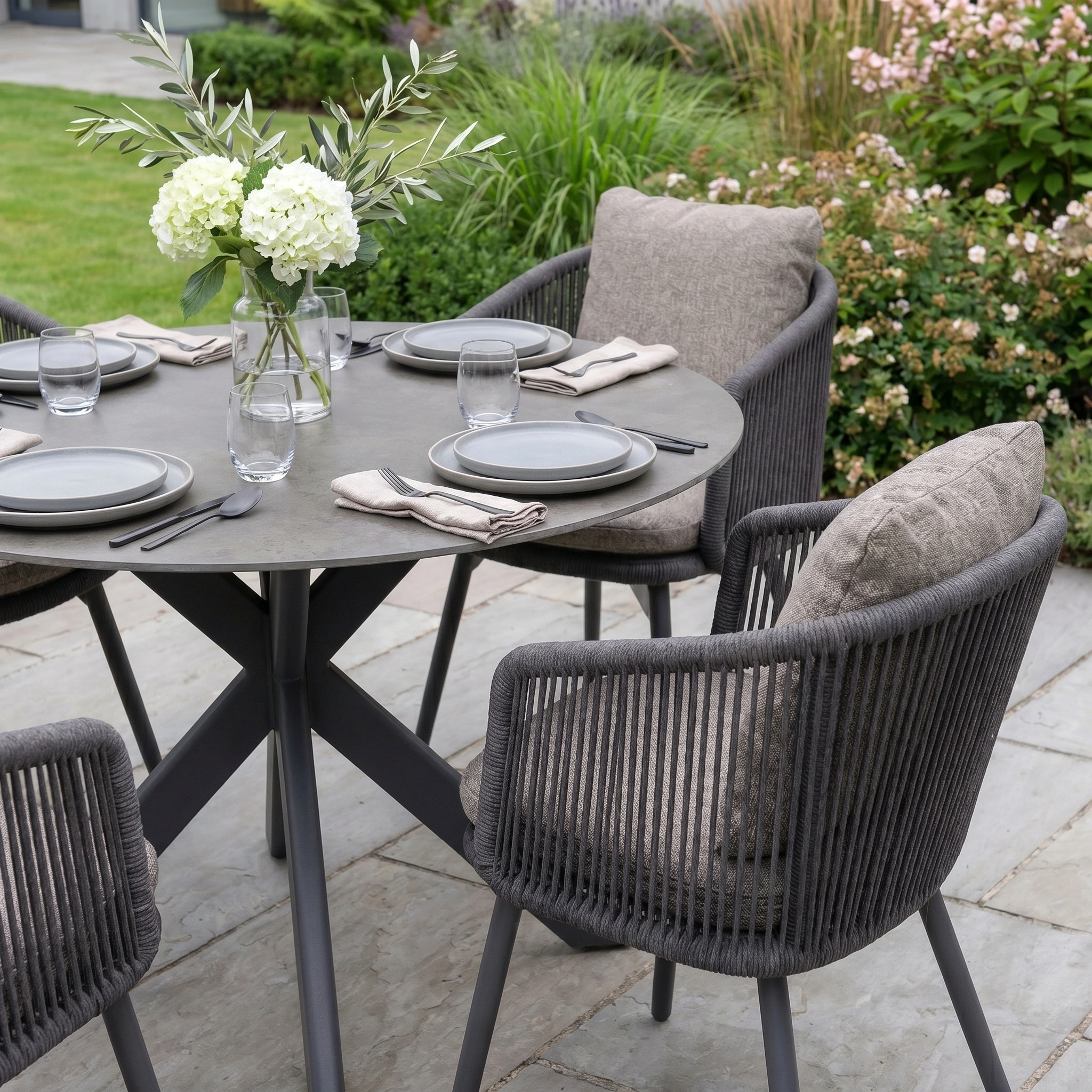The Theia 4 Seat Round Rope Dining Set with Albatross Table in Truffle features gray chairs, neutral plates, and a vase of white flowers—an elegant choice for memorable alfresco dining in your garden.