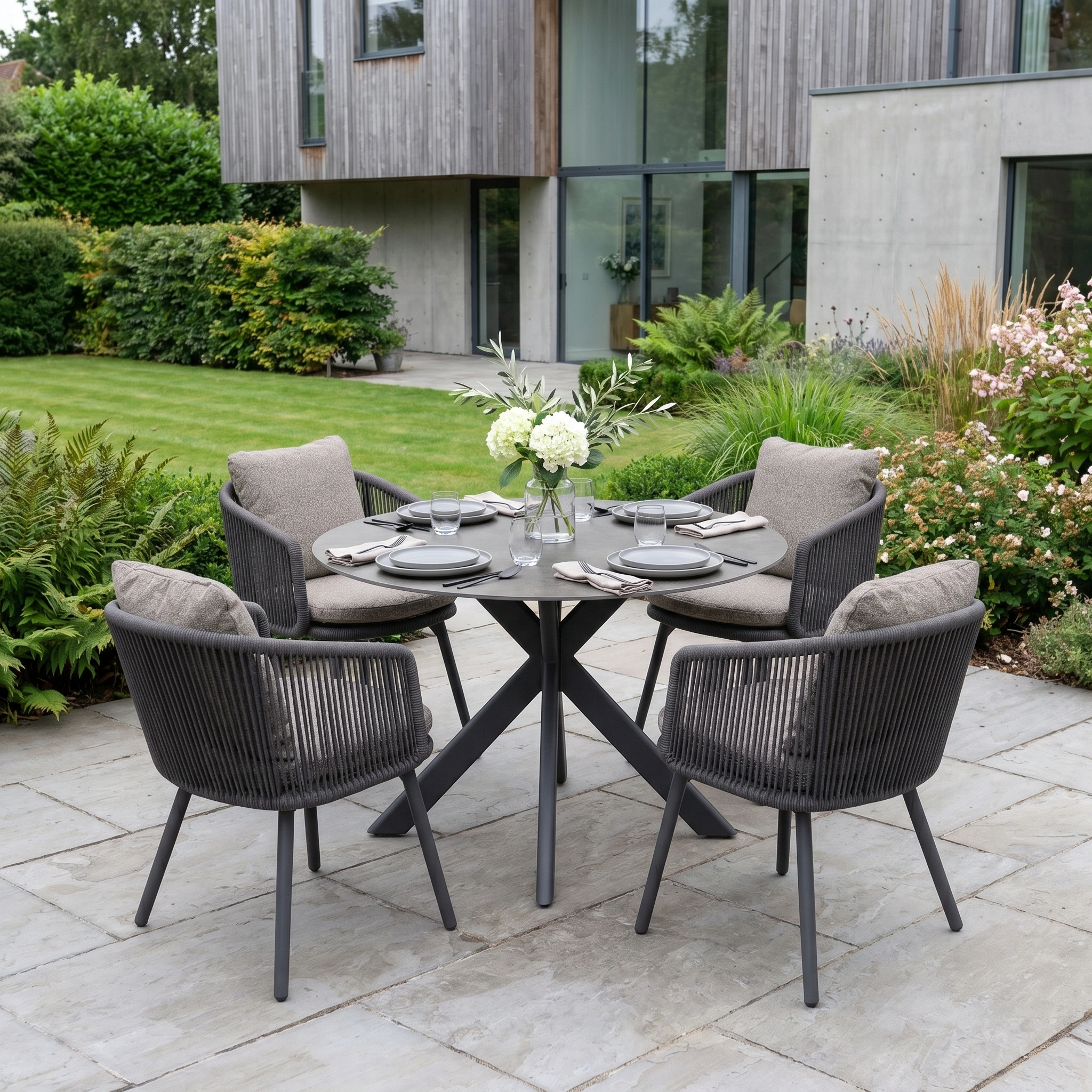 The Theia 4 Seat Round Rope Dining Set with Albatross Table in Truffle features four cushioned chairs and a round table, arranged on a stone patio for stylish outdoor dining.
