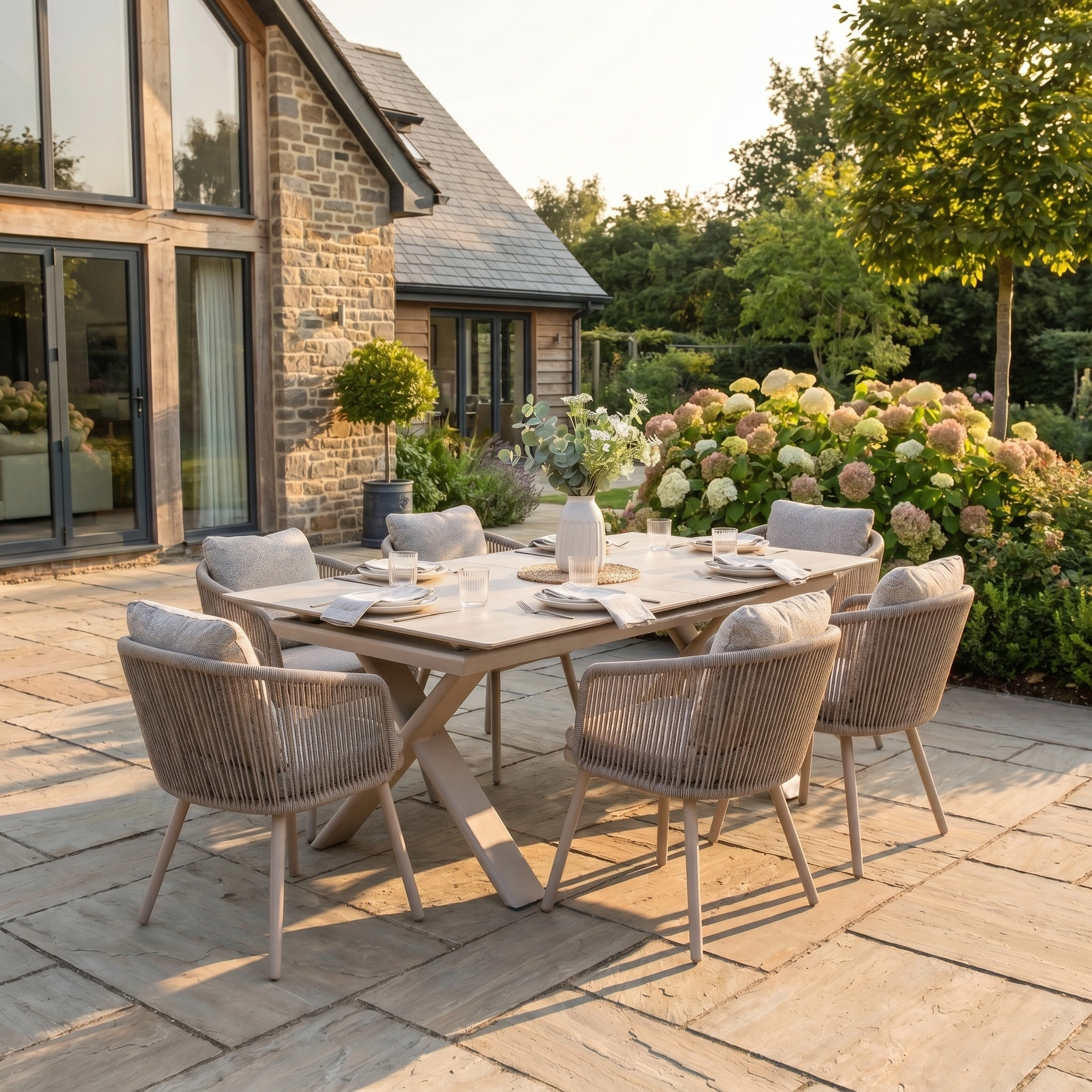 Modern outdoor dining set with the Theia 6 Seat Rope Dining Set and Albatross Extending Table in Oatmeal, shown on a stone patio amid lush greenery and flowers by a charming stone house.