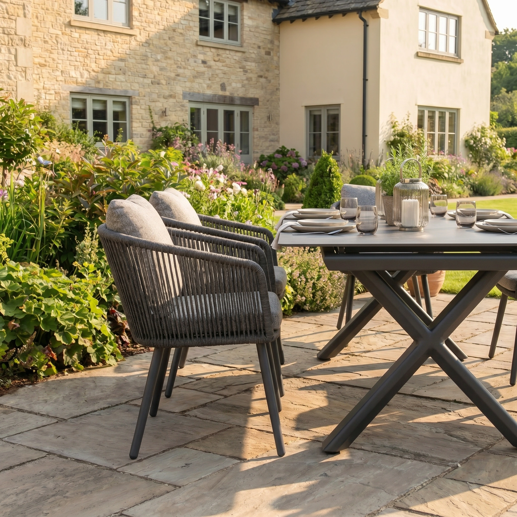 Enjoy stylish garden dining with the Theia 6 Seat Rope Dining Set featuring an Albatross Extending Table in Truffle, ideal for outdoor gatherings on a stone patio surrounded by lush gardens and a charming stone house.