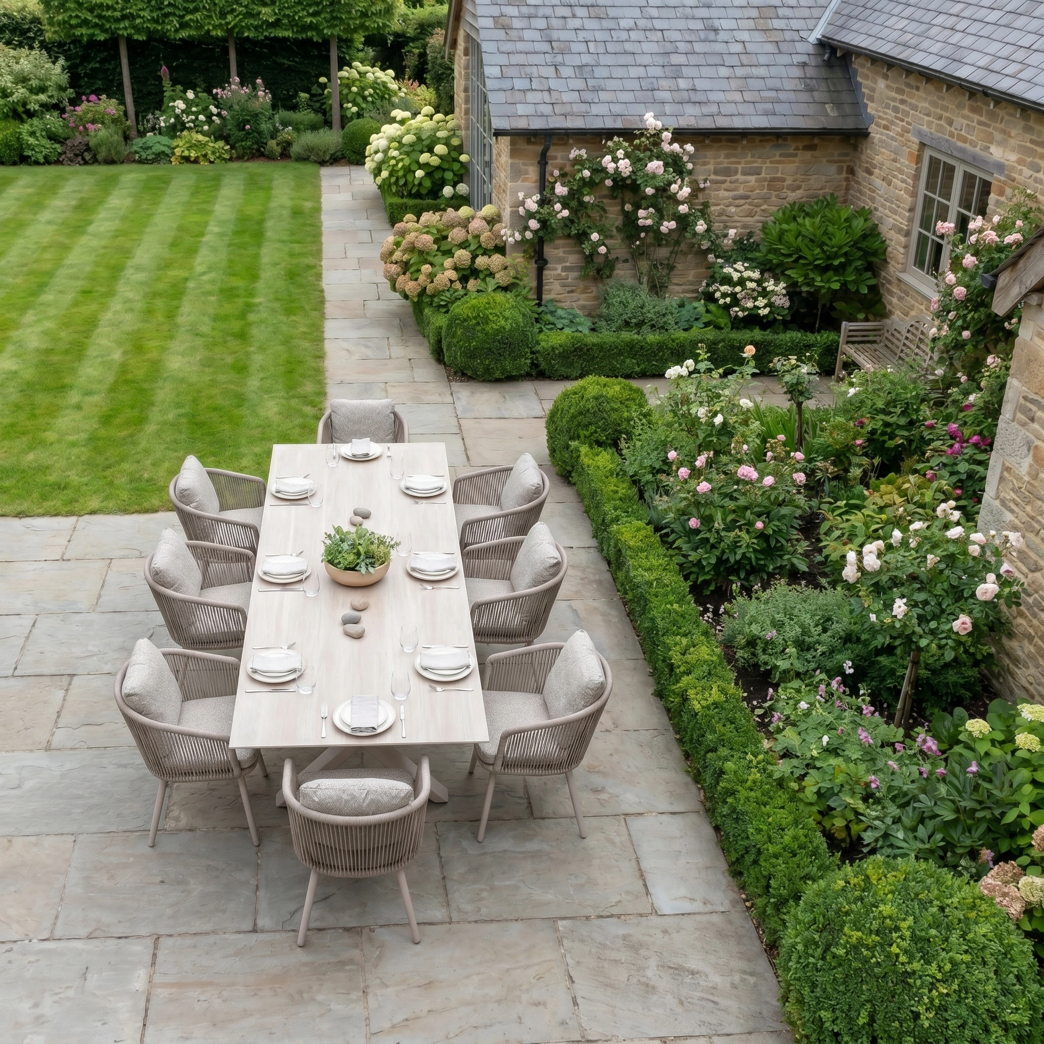 The Theia 8 Seat Rope Dining Set with Albatross Extending Table in Oatmeal stands beside a green garden and manicured lawn by a stone house, creating the ideal outdoor dining area.