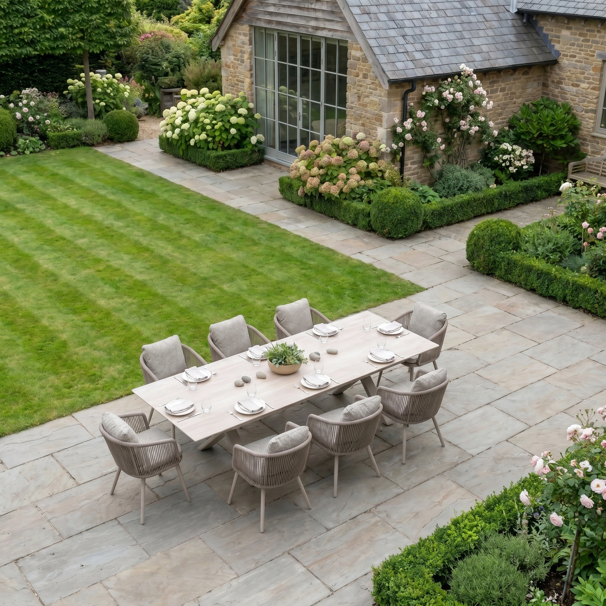 The Theia 8 Seat Rope Dining Set with Albatross Extending Table in Oatmeal is arranged on a stone patio in a well-kept garden.