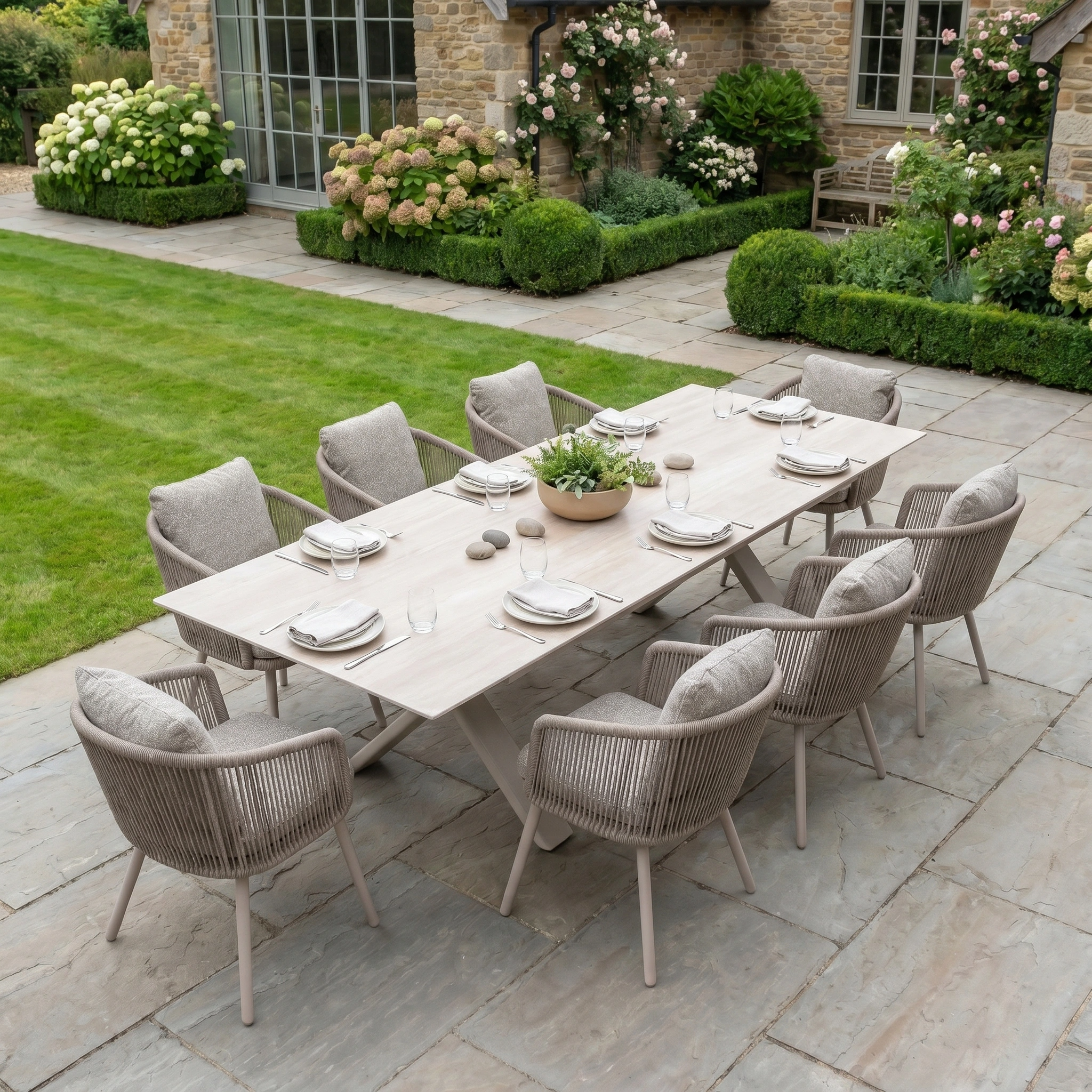 The Theia 8 Seat Rope Dining Set with Albatross Extending Table in Oatmeal is arranged for dining on a stone patio amid lush garden surroundings.