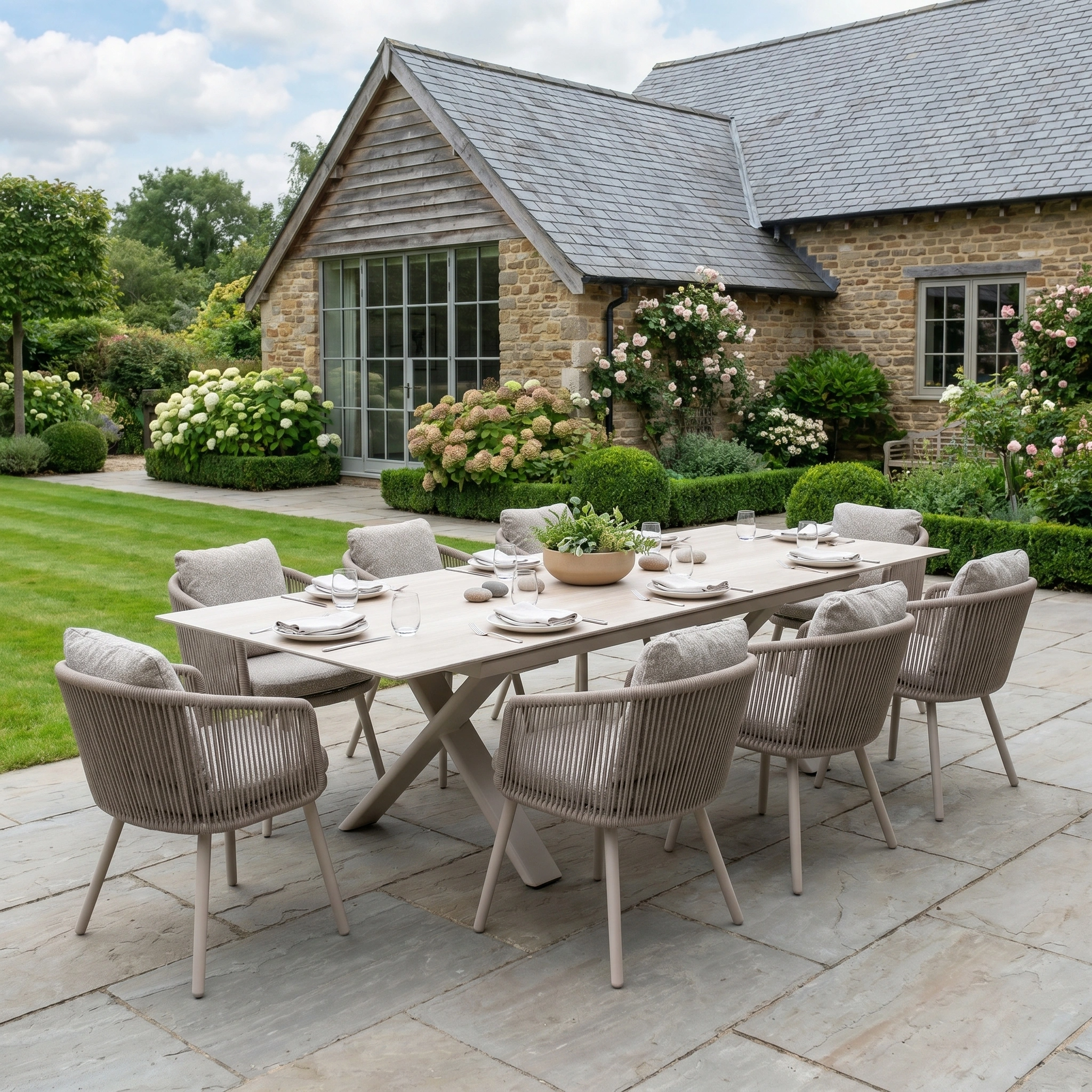 The Theia 8 Seat Rope Dining Set with Albatross Extending Table in Oatmeal is displayed on a stone patio in a garden next to a rustic house.