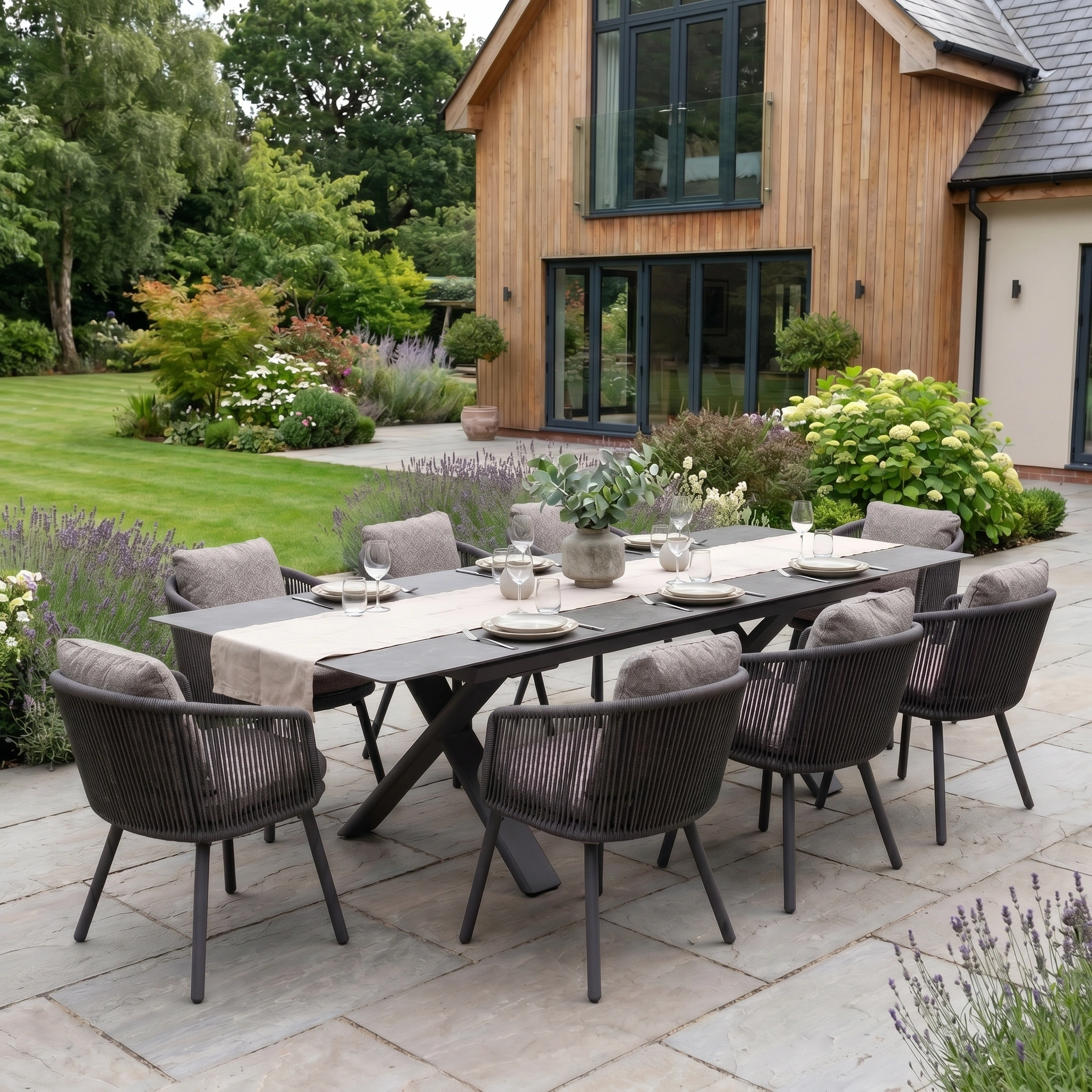 Theia 8 Seat Rope Dining Set with Albatross Extending Table in Truffle, stylishly arranged on a patio in a lush garden outside a wooden house.