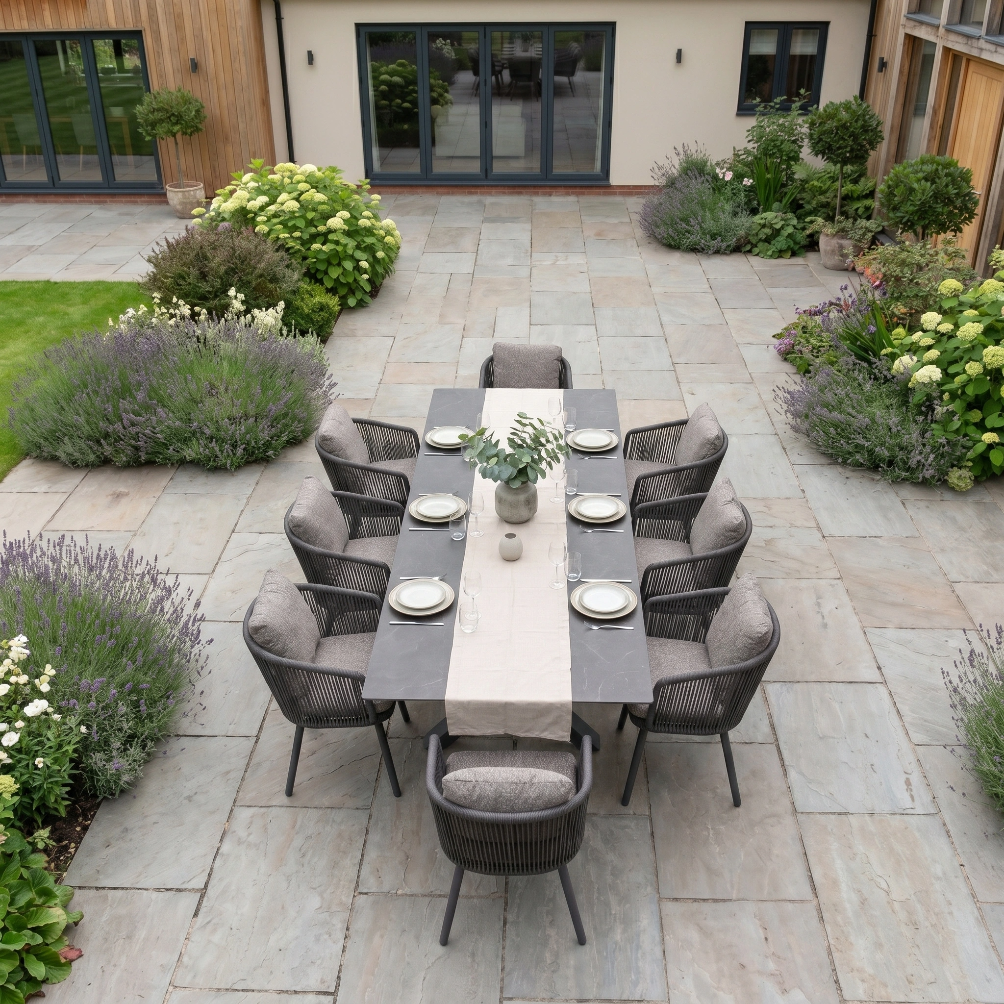 The Theia 8 Seat Rope Dining Set with Albatross Extending Table in Truffle is showcased on a stone patio, surrounded by lush gardens and a modern house exterior.