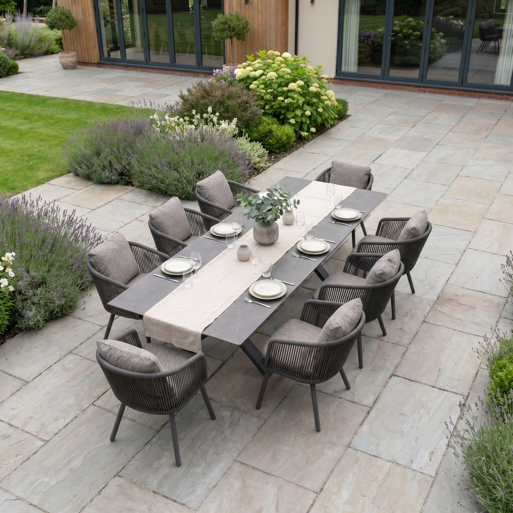 Theia 8 Seat Rope Dining Set with Albatross Extending Table in Truffle on a stone patio, surrounded by green plants—an inviting piece of outdoor garden furniture perfect for stylish entertaining.