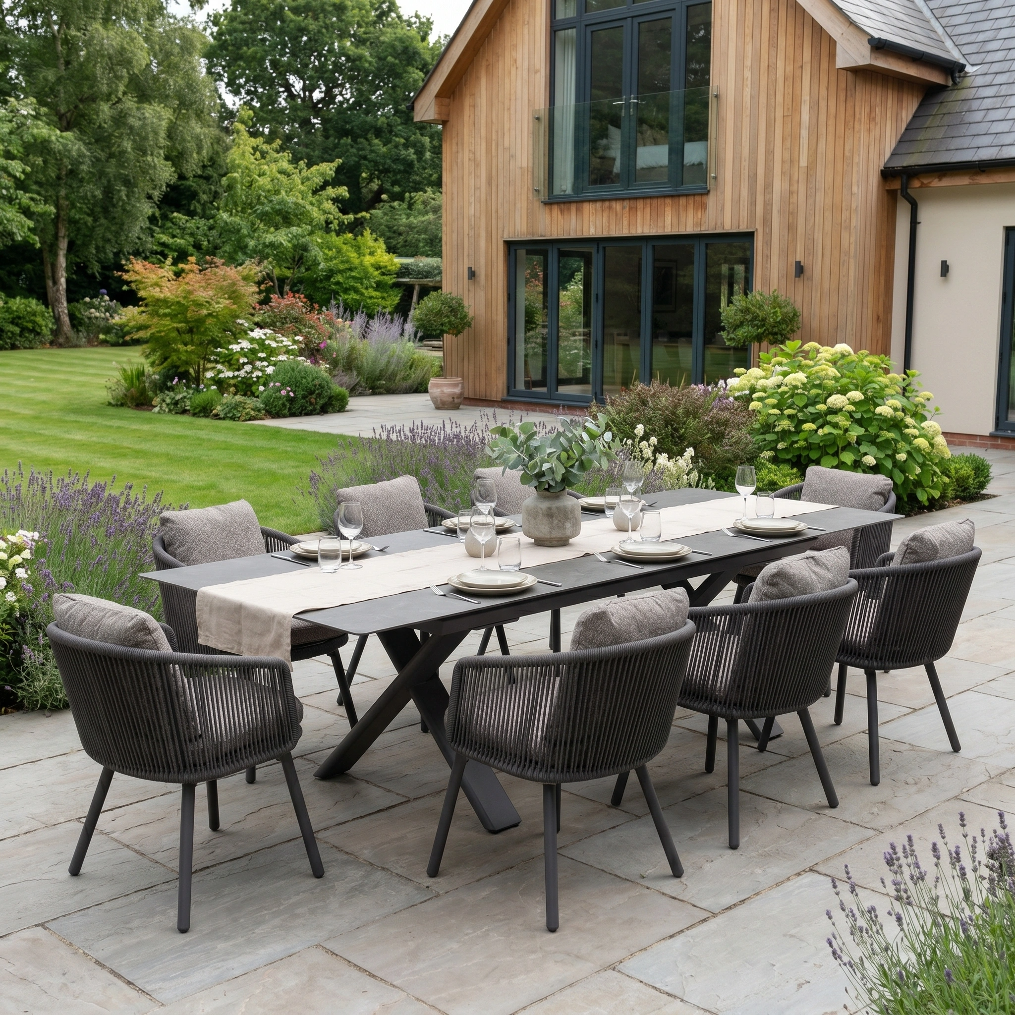 The Theia 8 Seat Rope Dining Set with Albatross Extending Table in Truffle is arranged on a stone patio in a beautifully landscaped garden, ready for an elegant al fresco meal.