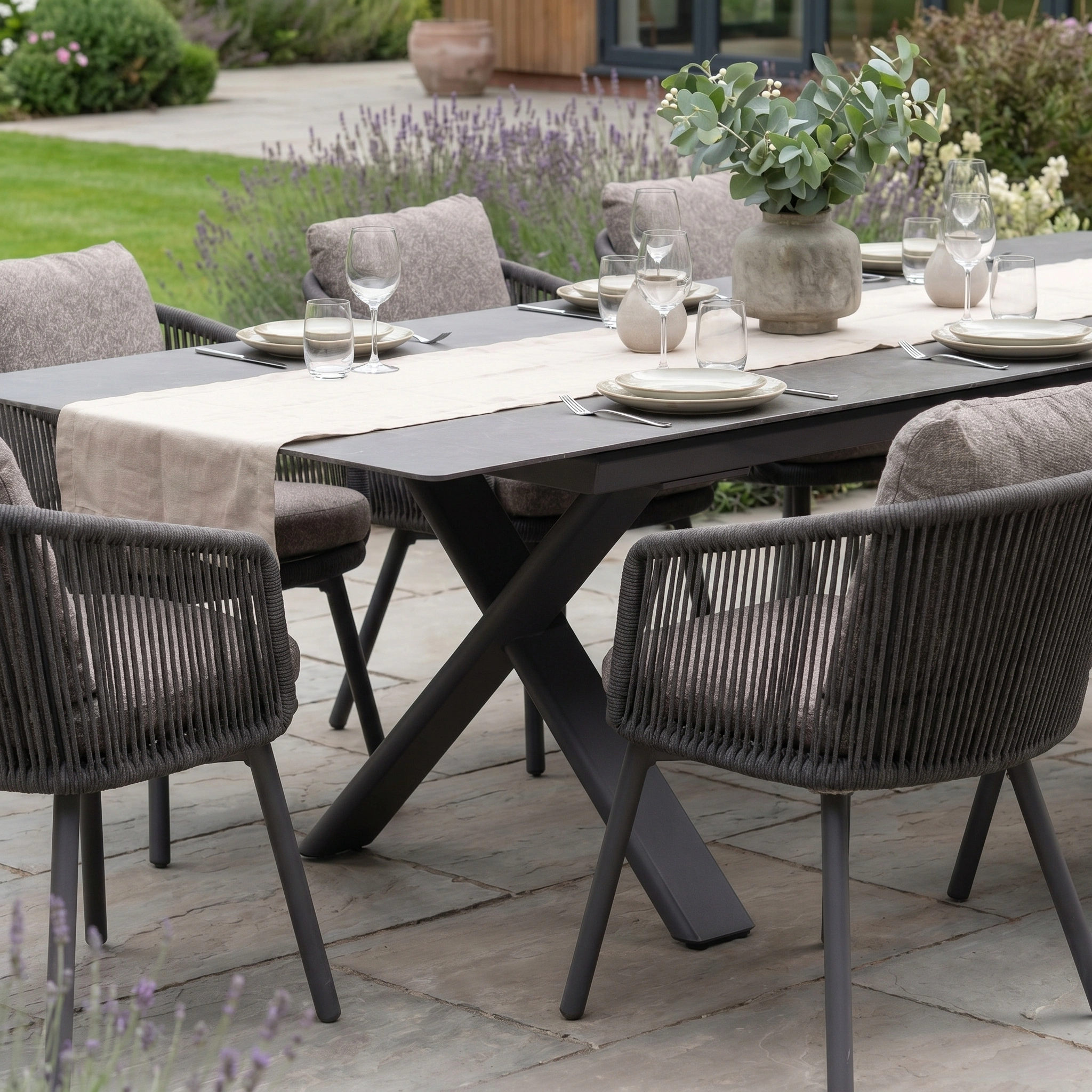 Theia 8 Seat Rope Dining Set with Albatross Extending Table in Truffle—ideal for gatherings, featuring space for up to eight on a stylish truffle stone patio, surrounded by greenery, with included plates, glasses, and vase.