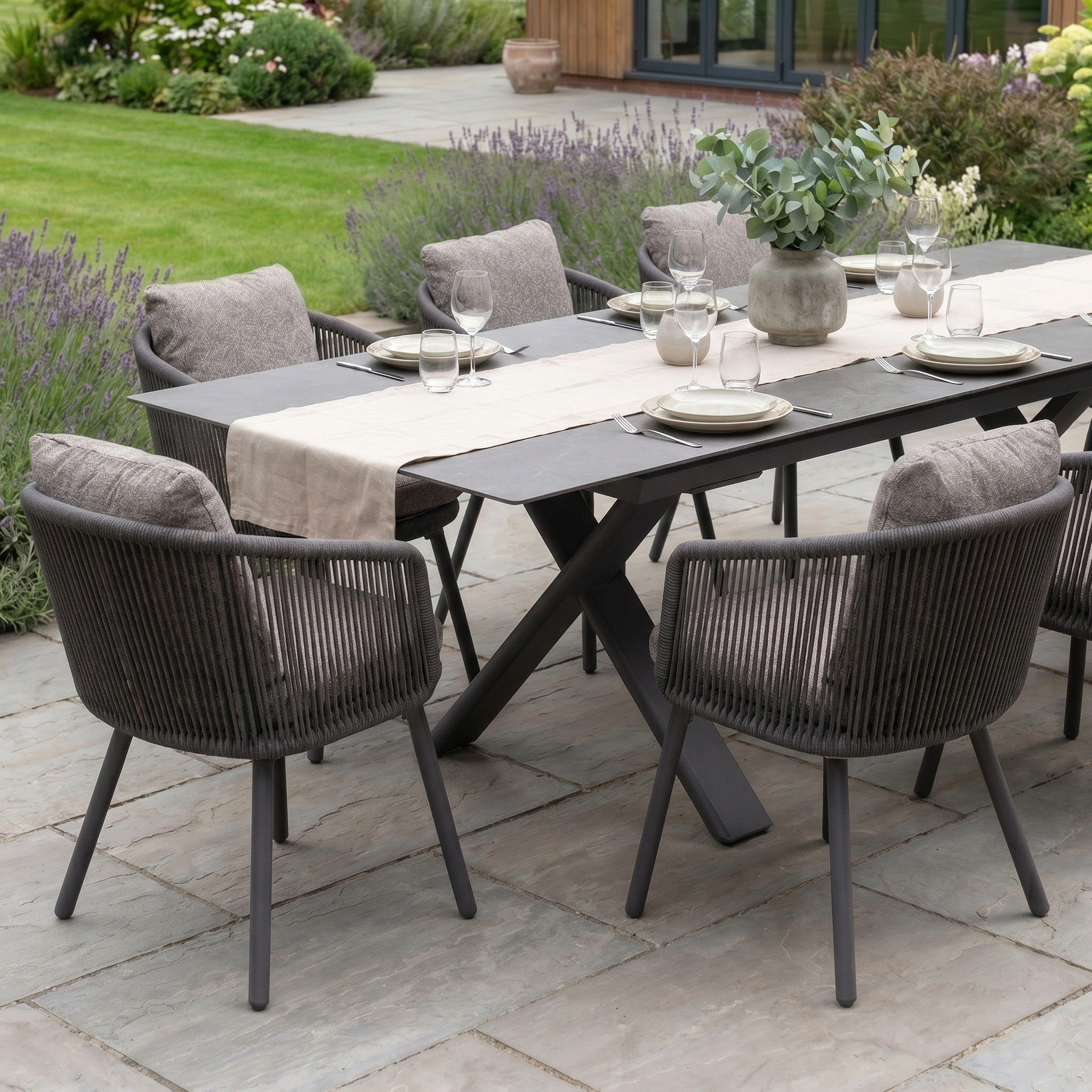 Theia 8 Seat Rope Dining Set with Albatross Extending Table in Truffle, arranged for dining with eight cushioned chairs on a stone patio amidst a lush garden.
