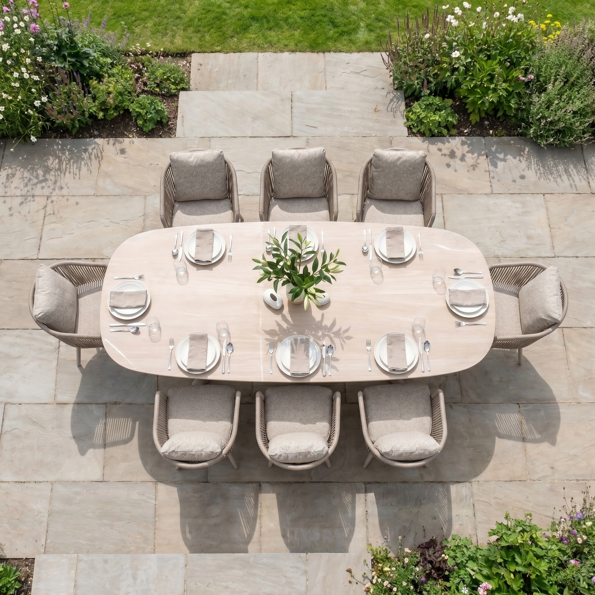 The Theia 8 Seat Oval Rope Dining Set with Albatross Extending Table in Oatmeal is styled with plates, glasses, and a vase on a stone patio for outdoor dining.