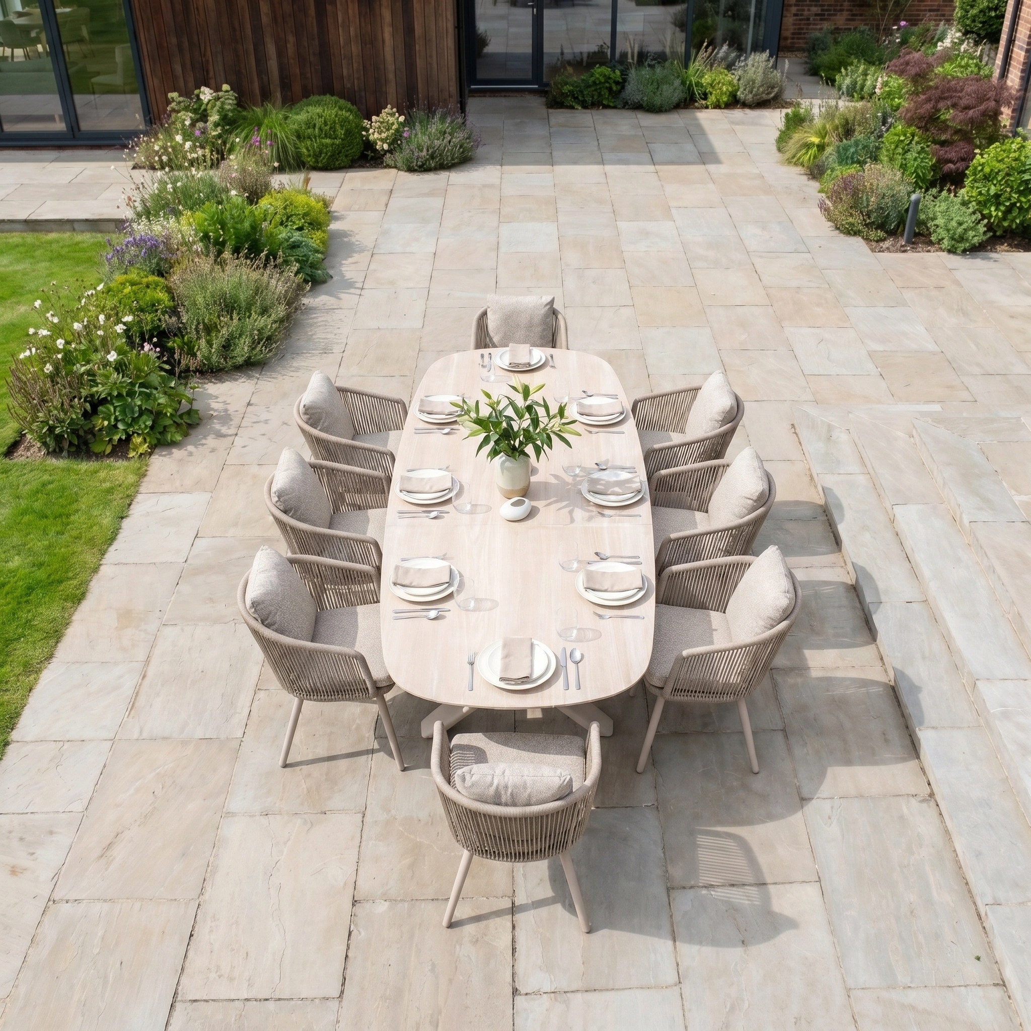 The Theia 8 Seat Oval Rope Dining Set with Albatross Extending Table in Oatmeal features stylish rope dining chairs, perfect for outdoor meals on a stone patio surrounded by lush greenery.
