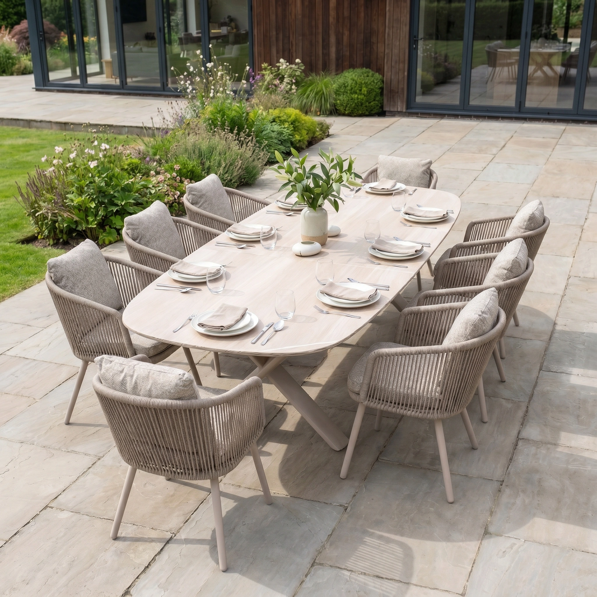 The Theia 8 Seat Oval Rope Dining Set with Albatross Extending Table in Oatmeal features cushioned rope chairs and a stylish table, perfect for outdoor dining on a stone patio next to a beautiful garden.