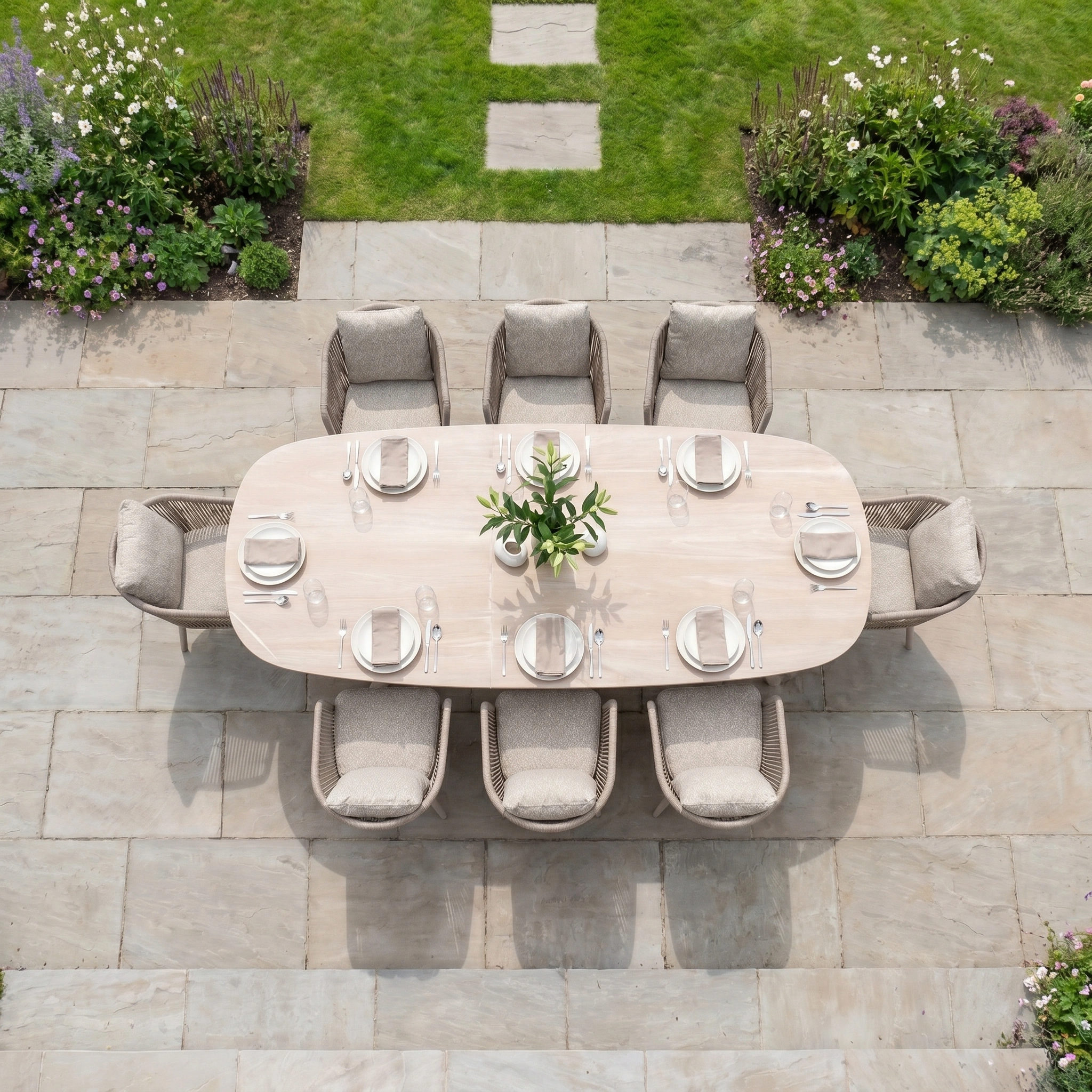 The Theia 8 Seat Oval Rope Dining Set with Albatross Extending Table in Oatmeal is displayed on a stone patio, surrounded by lush garden greenery and flowers.