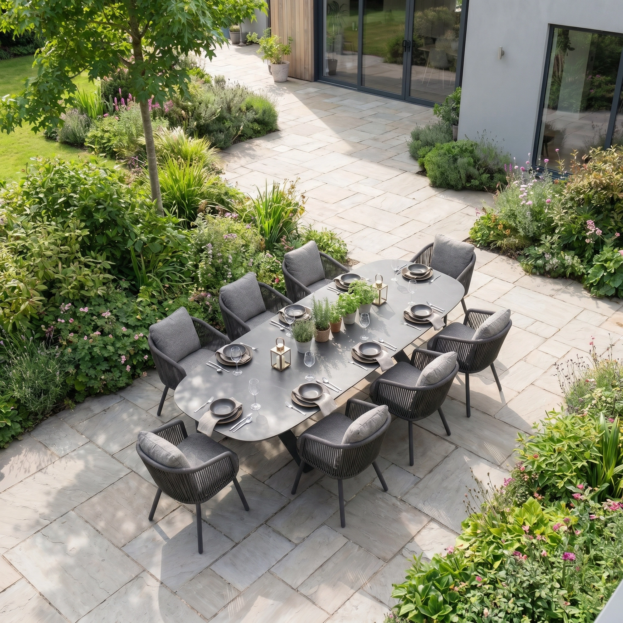 The Theia 8 Seat Oval Rope Dining Set with Albatross Extending Table in Truffle sits on a patio, its elegant grey chairs surrounded by lush garden greenery.