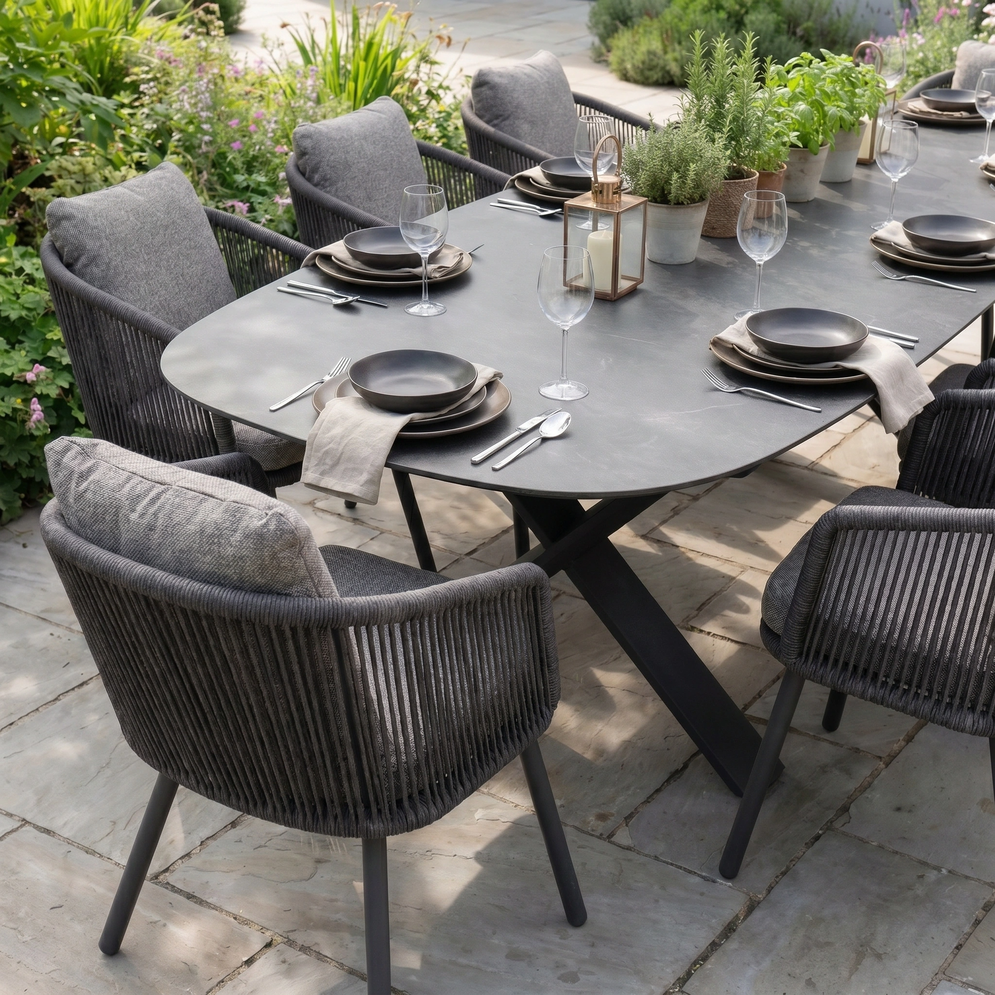 Host six with the Theia 8 Seat Oval Rope Dining Set featuring the Albatross Extending Table in Truffle, gray chairs, black plates, wine glasses, and potted plants as a centerpiece.