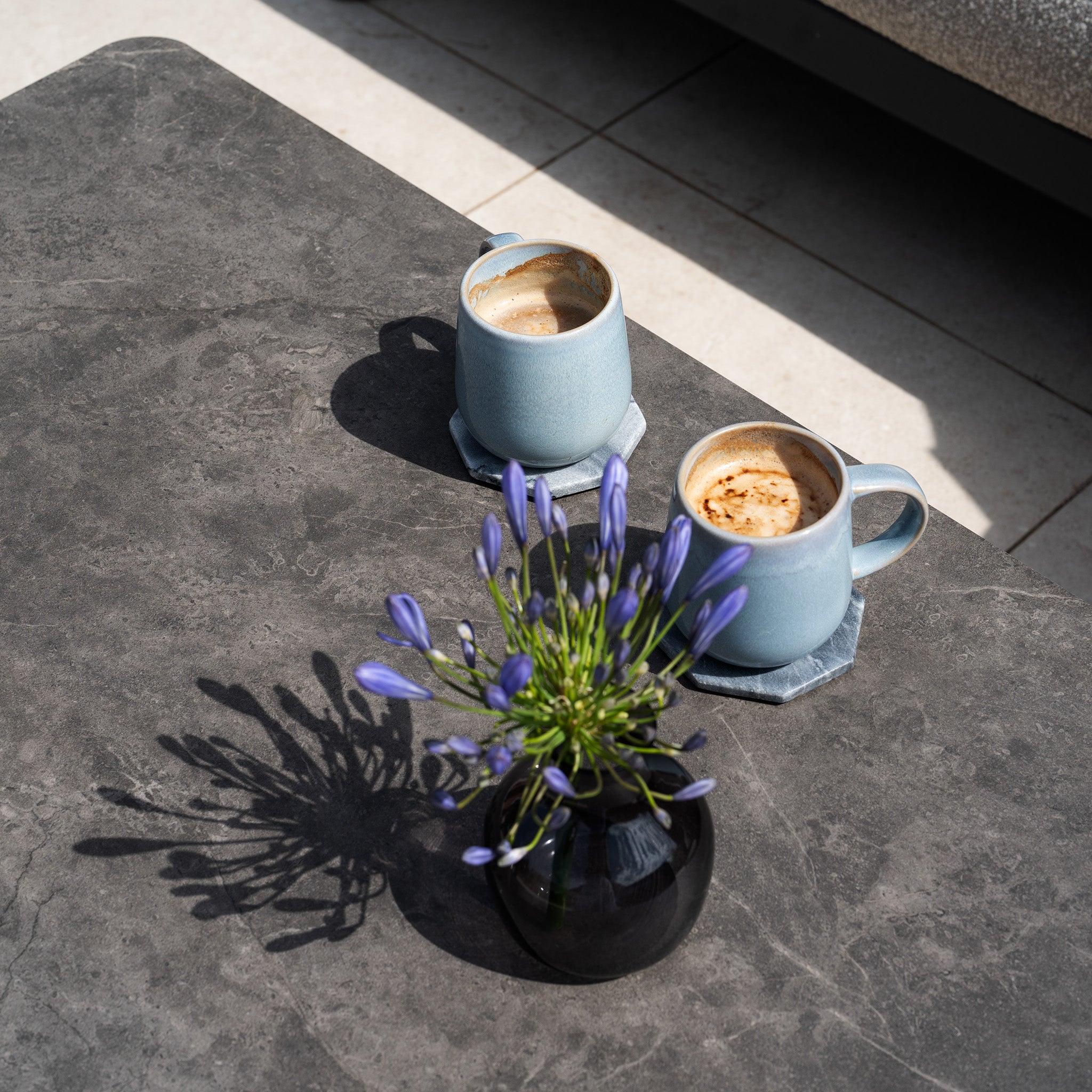 Two blue mugs of coffee rest on a gray table beside a black vase filled with purple flowers, while the Theia 3 Seat Sofa Set in Truffle completes the modern outdoor furniture arrangement.