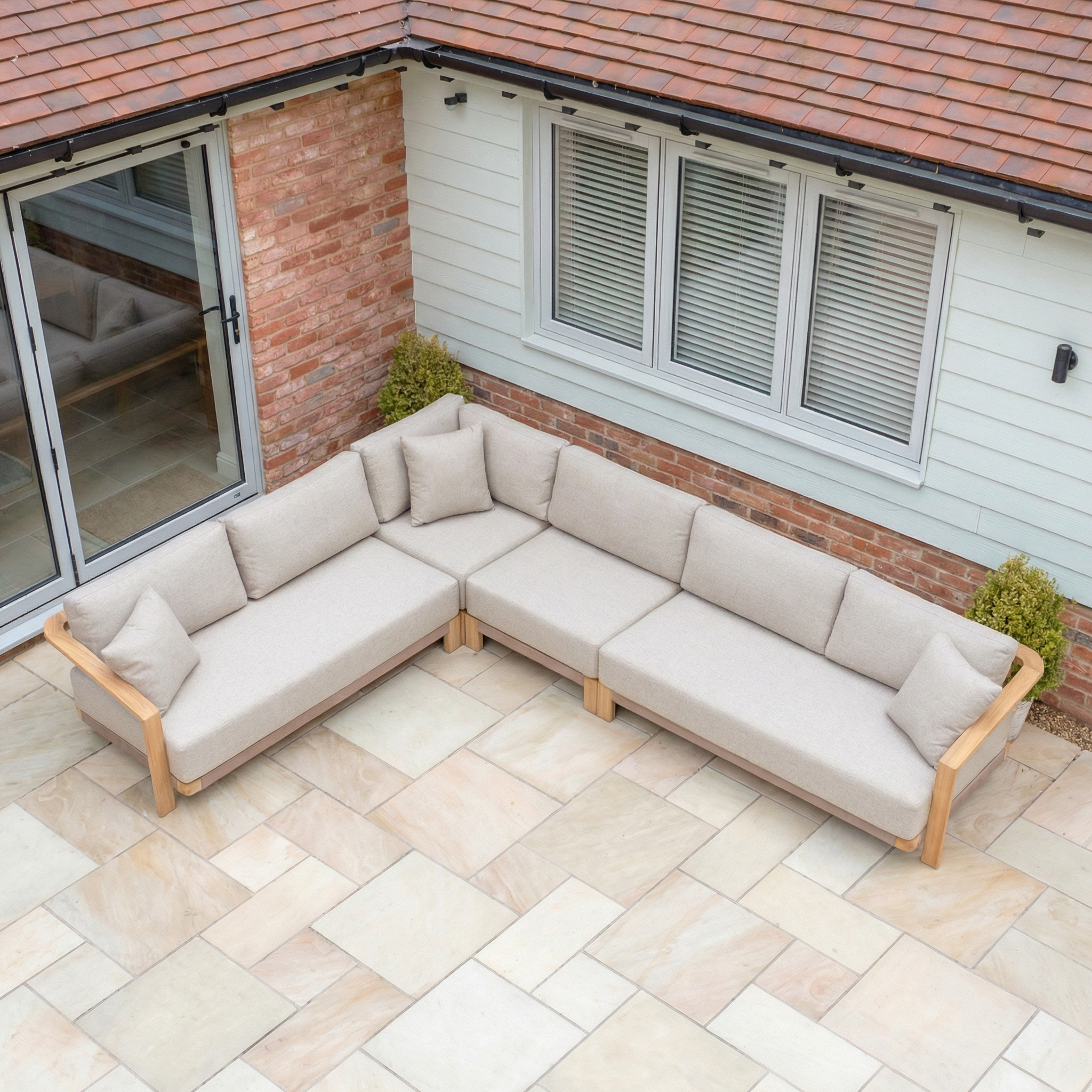 The Tivoli Large Corner Sofa in Fawn (Ex Display), an L-shaped outdoor sofa with a sturdy wooden frame, sits on a stone patio by large windows and is paired with a marble coffee table.