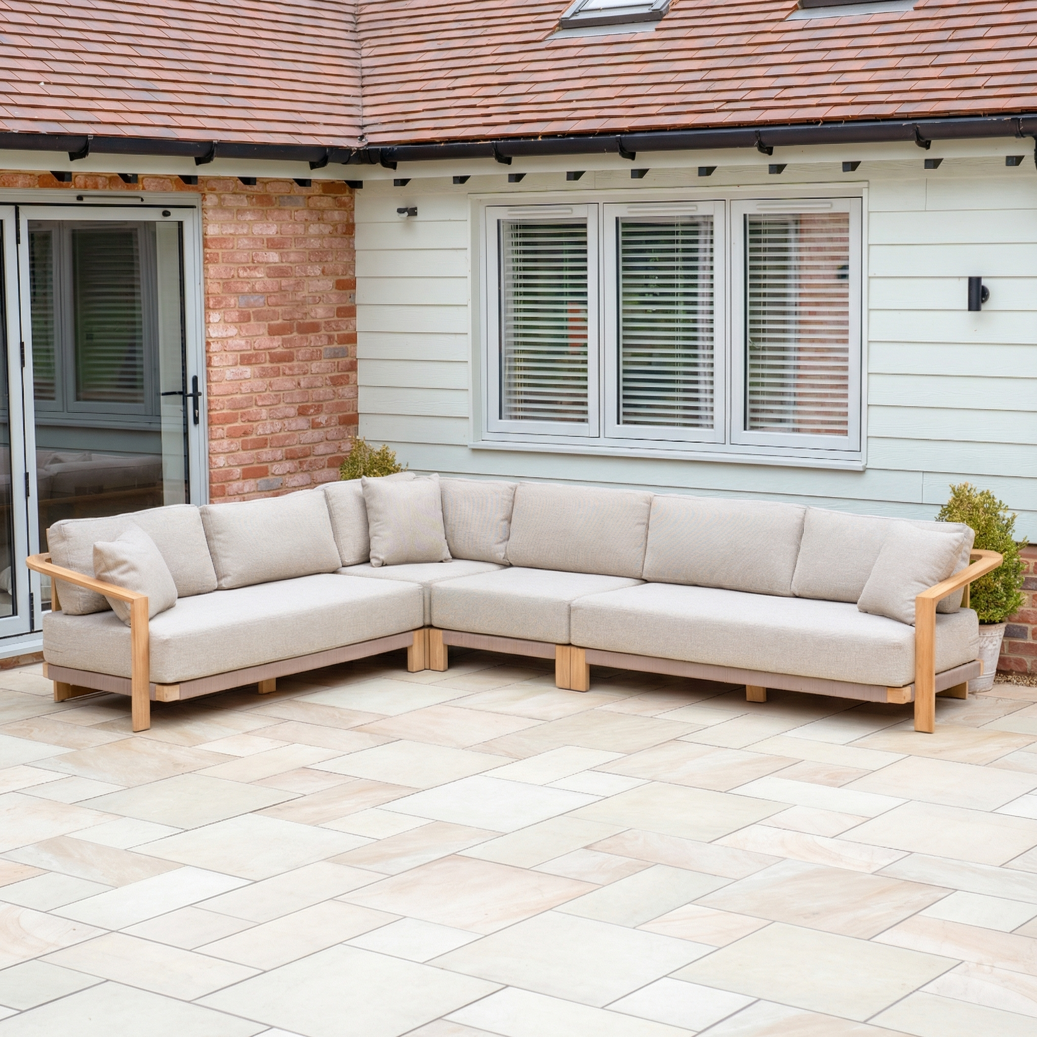 The Tivoli Large Corner Sofa in Fawn (Ex Display) sits on a tiled patio outside a house with windows and a glass door, paired with a marble coffee table—ideal for stylish outdoor lounging.