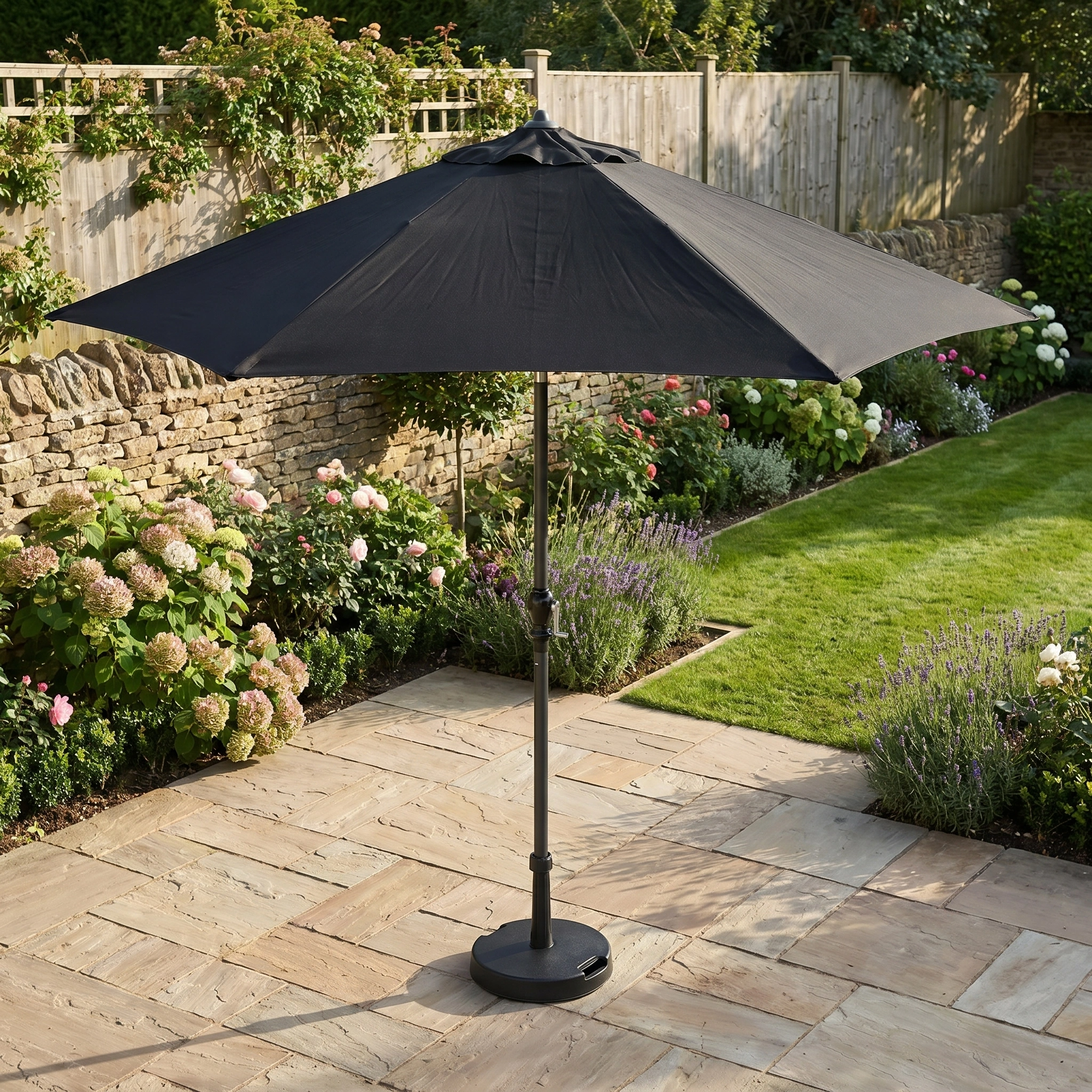 The Triton 2.5m Round Aluminium Parasol in Black offers stylish UV protection on a stone patio, enhancing your landscaped garden among vibrant flowers and lush grass.