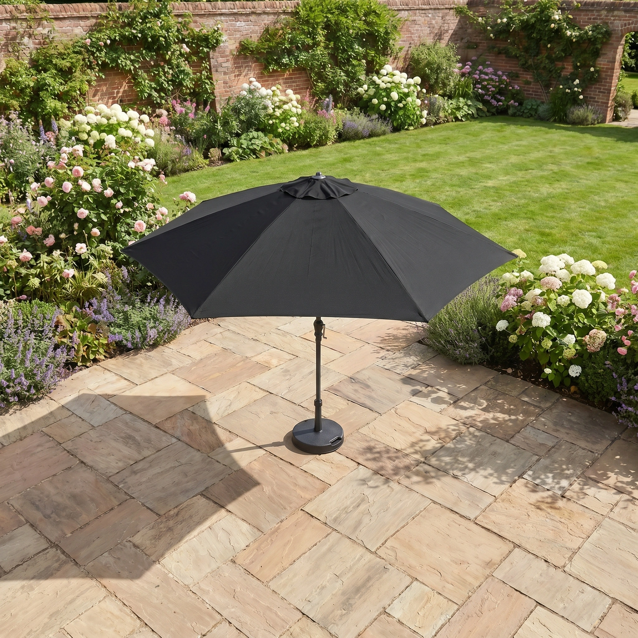 The Triton 2.5m Round Aluminium Parasol in Black stands on a stone terrace by a green lawn and flowering plants, providing stylish UV protection for your garden.
