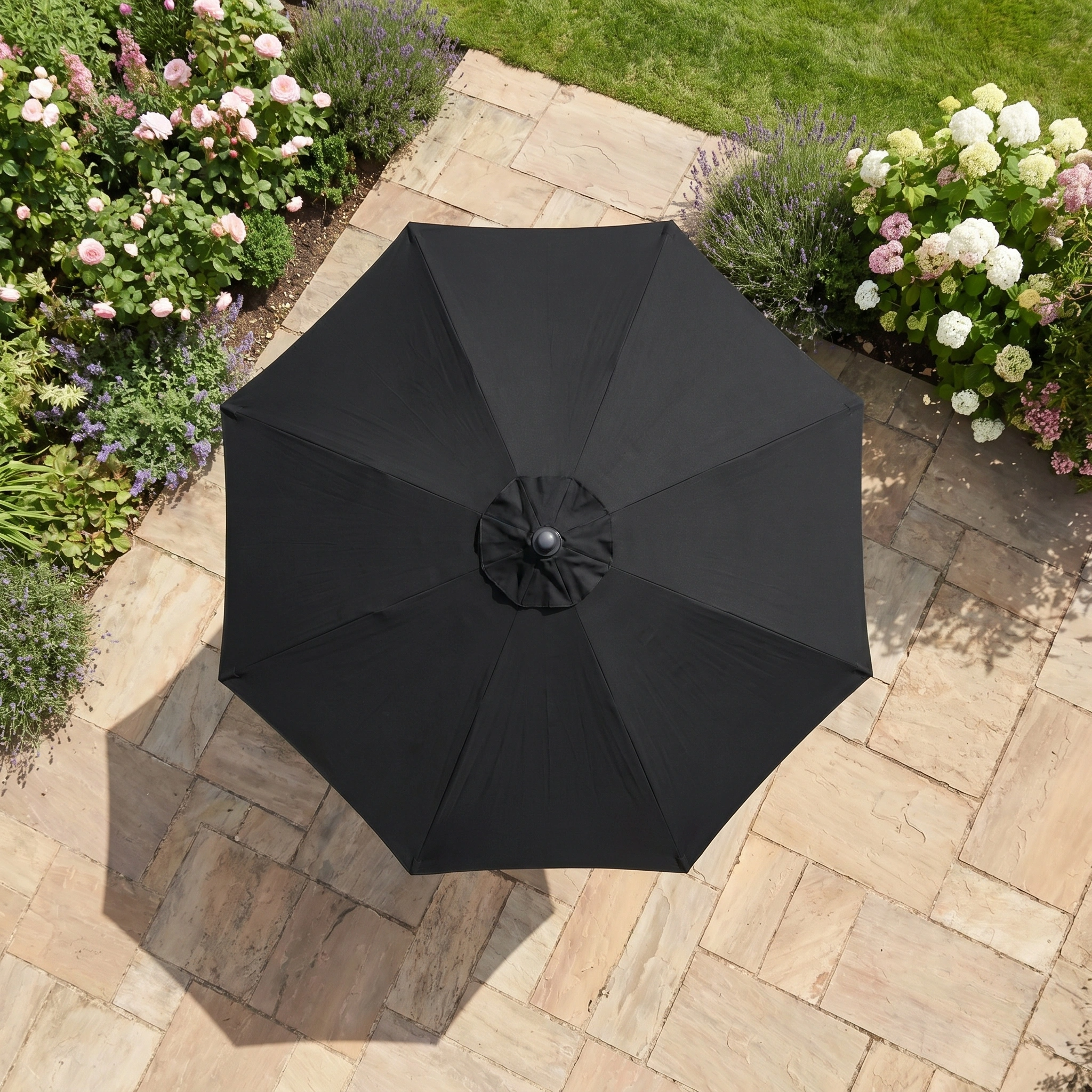 The Triton 2.5m Round Aluminium Parasol in Black, seen from above among stone tiles and blooming flowers, adds stylish UV protection to your outdoor space.