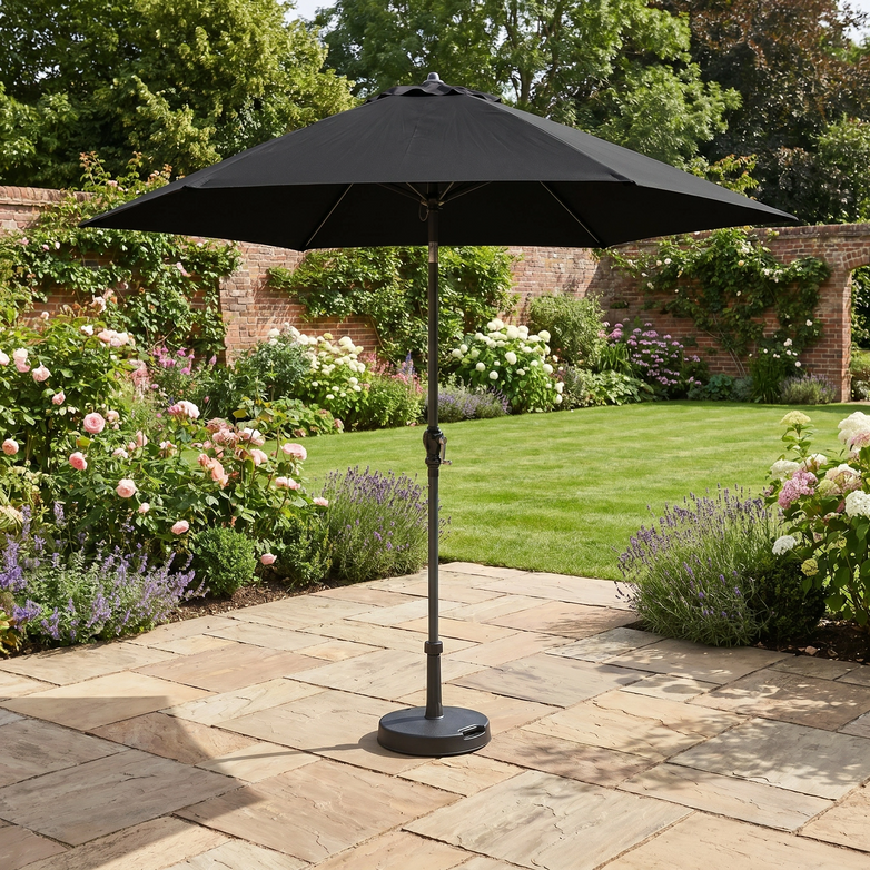 The Triton 2.5m Round Aluminium Parasol in Black stands on a stone terrace in a flower-filled garden with a green lawn, providing stylish shade and UV protection.