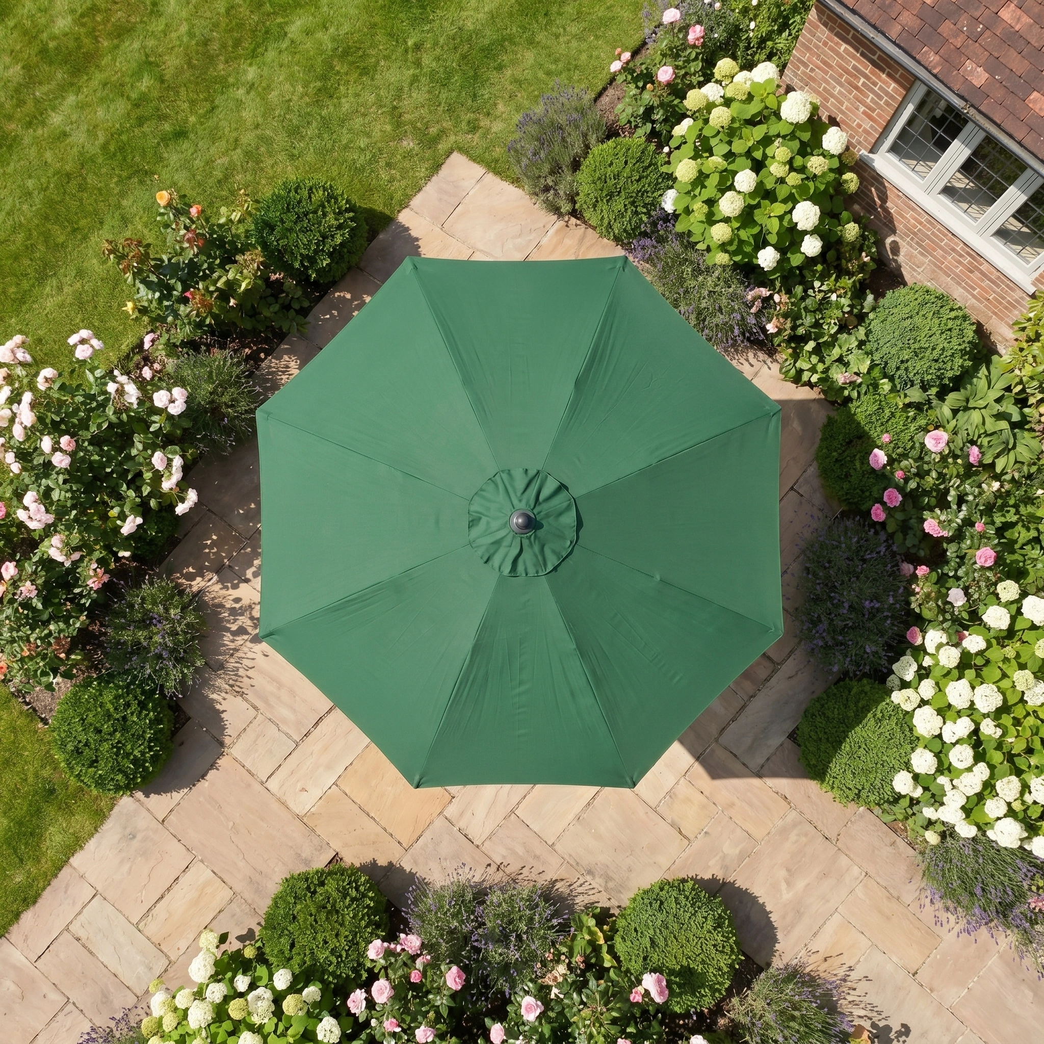 The Triton 2.5m Round Aluminium Parasol in Green stands on a stone terrace amid blooming flowers and green bushes, providing beauty and shade to your outdoor space.