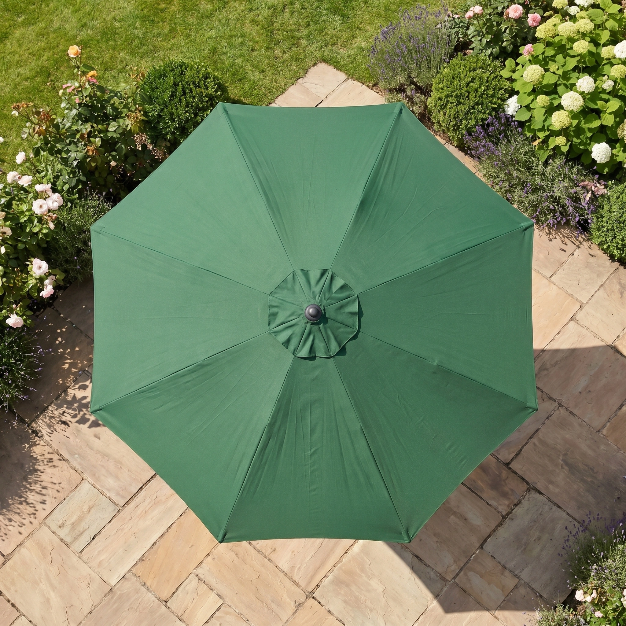 The Triton 2.5m Round Aluminium Parasol in Green provides reliable UV protection for your outdoor space, pictured from above on a stone patio bordered by flowers and grass.