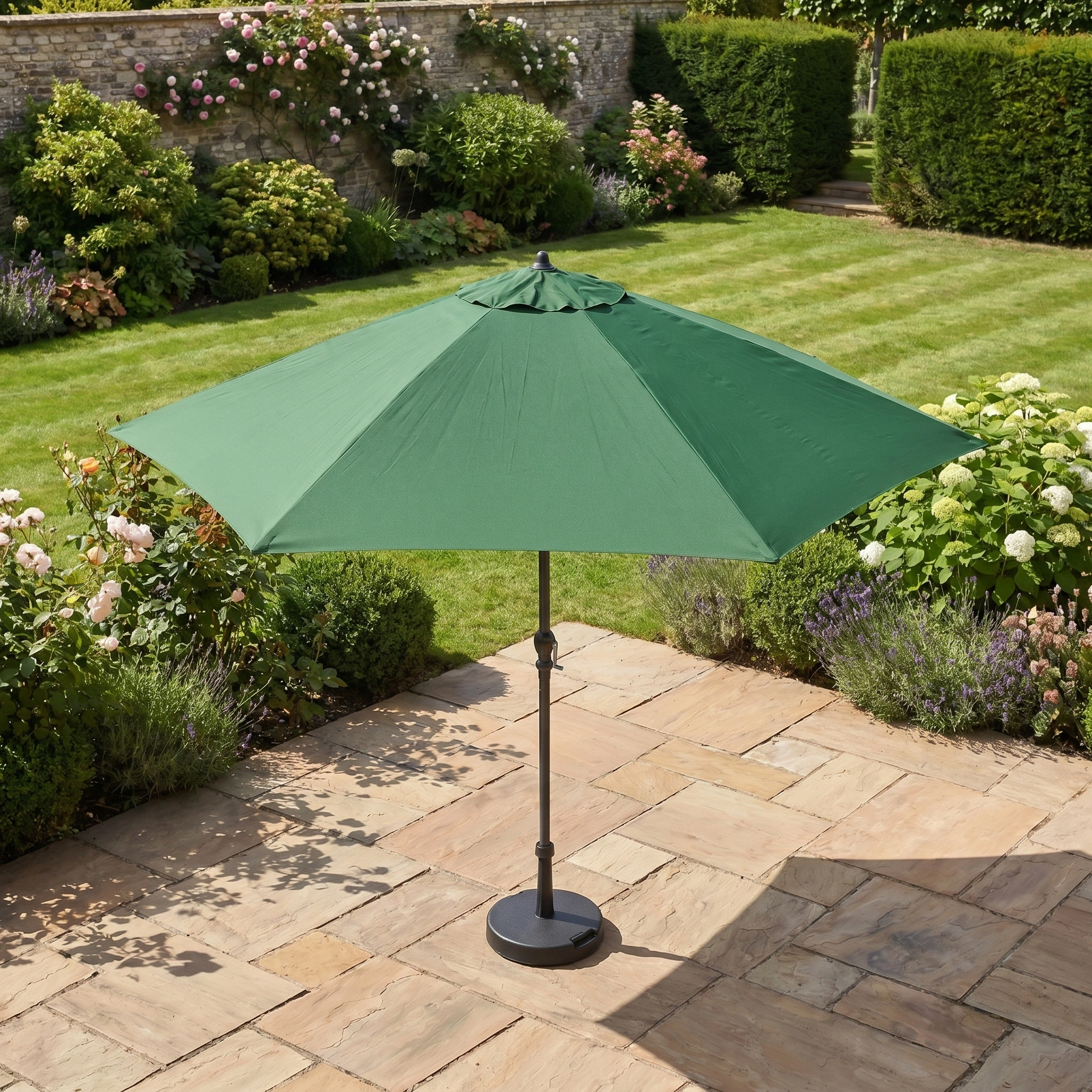 The Triton 2.5m Round Aluminium Parasol in Green stands open on a stone patio among flowers and manicured lawn, adding style and UV protection to the garden.